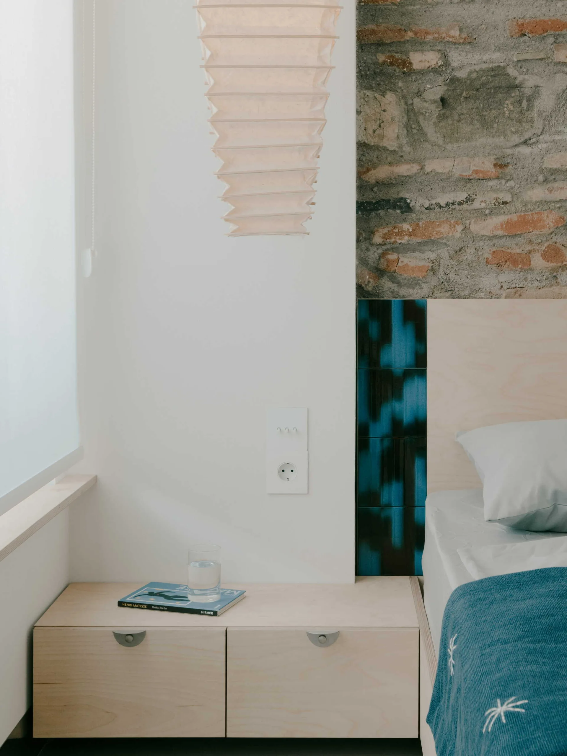 A bedroom corner with a light wood nightstand, a pleated paper pendant light, a white wall, and a vertical stripe of blue tiles next to exposed brick.