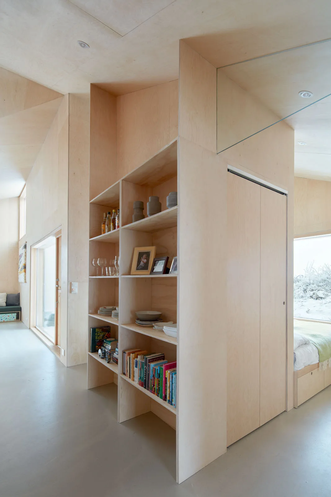 A minimalist light wood plywood interior featuring a custom floor-to-ceiling shelving partition that separates the hallway from an adjacent bedroom.