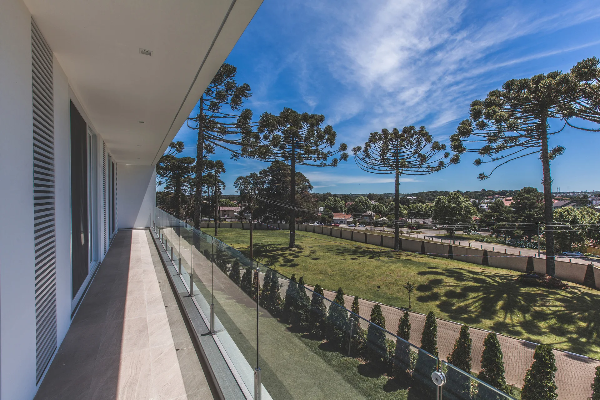 A long modern balcony features light grey stone-like floor tiles, a frameless glass railing, and a view of a grassy field with mature trees.