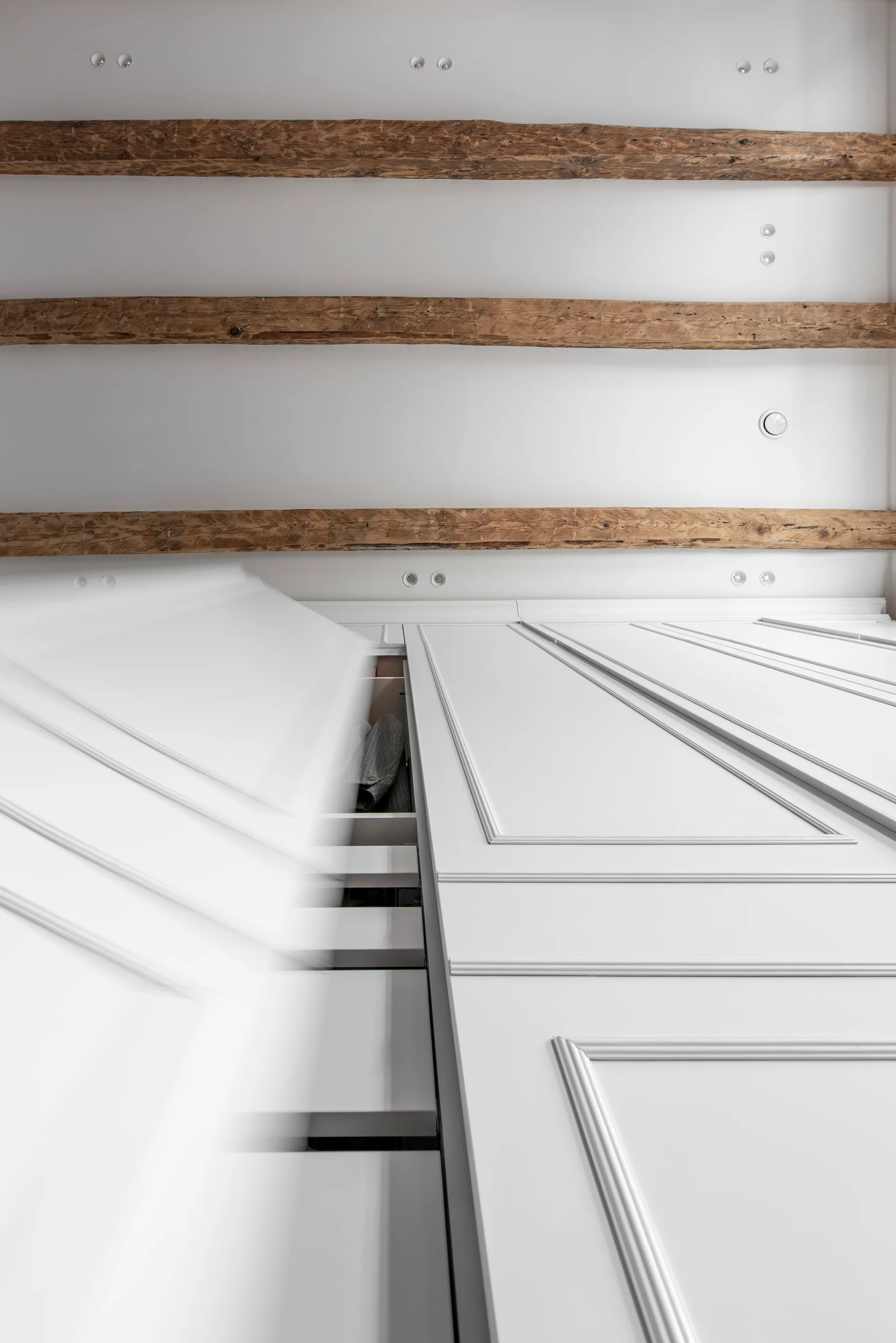 A low-angle shot of white custom paneled wardrobe doors opening under rustic wooden ceiling beams to reveal interior shelving.