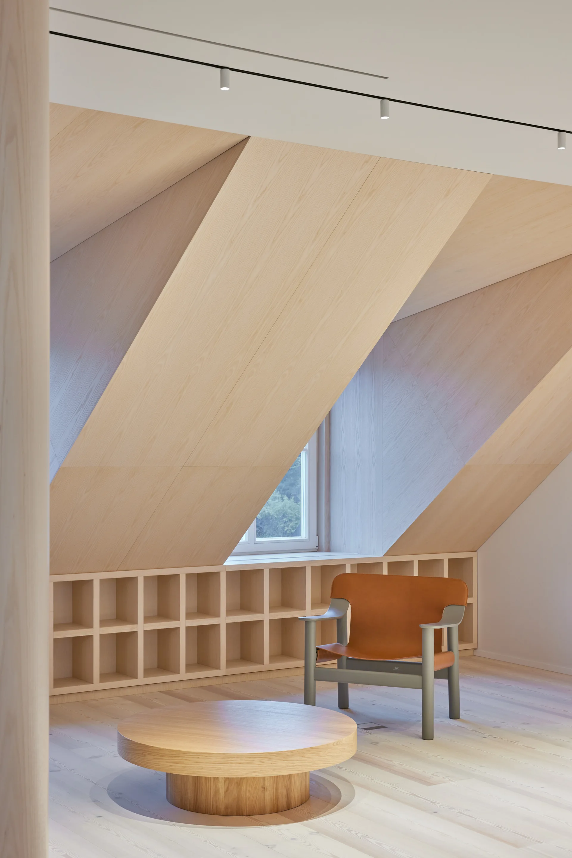 A minimalist attic room featuring slanted light wood paneled walls, a low wooden grid bookshelf under the window, and an orange leather armchair.