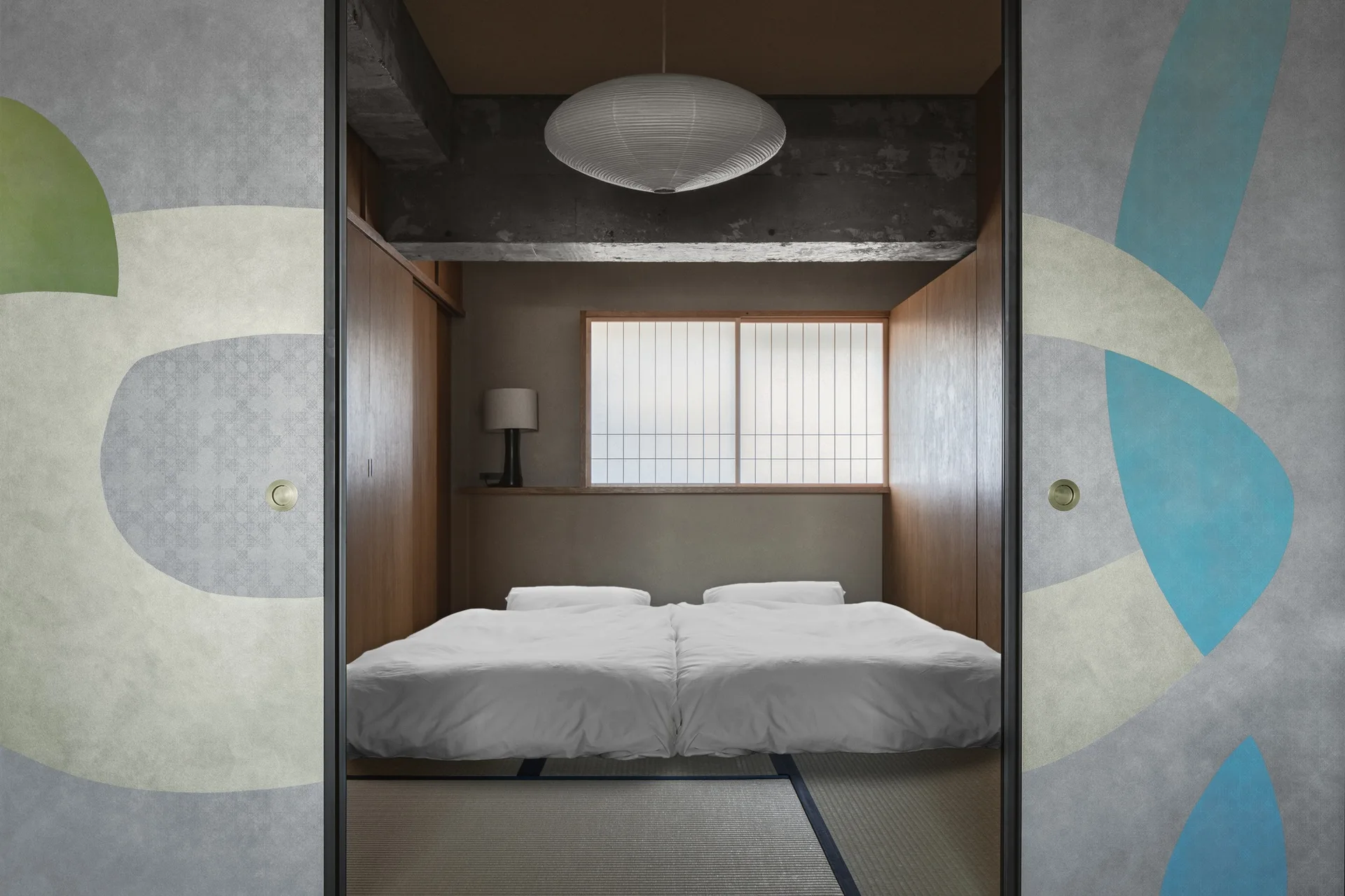 A minimalist bedroom with two beds on tatami mats, viewed through sliding doors detailed with abstract painted shapes.