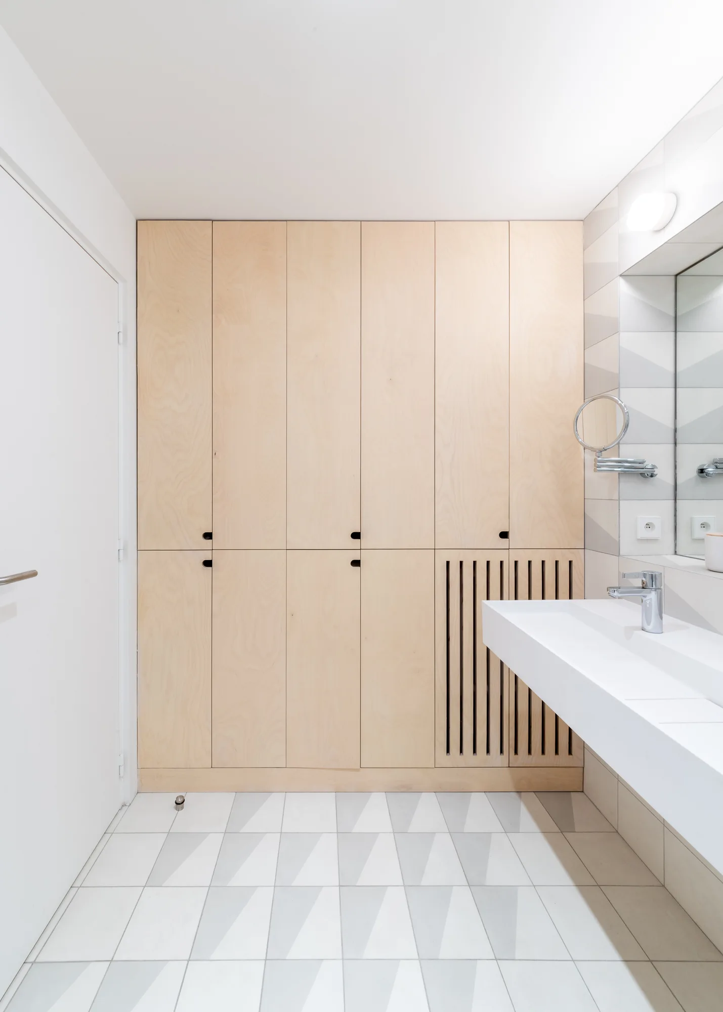A modern bathroom featuring full-height light wood cabinets, a floating white trough sink, and geometric grey and white triangular floor tiles.
