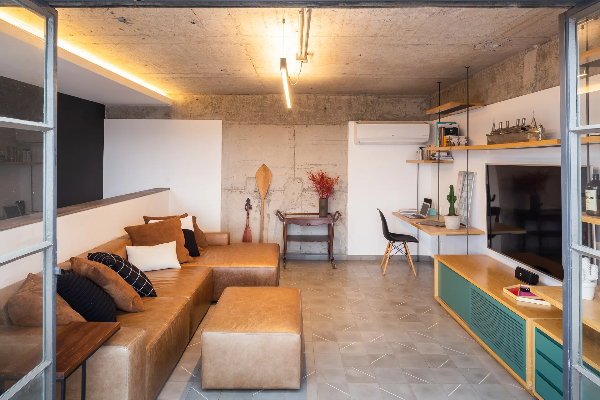 An industrial living room features a brown leather sectional sofa, a concrete ceiling, and a wall-mounted wooden desk with a black chair.