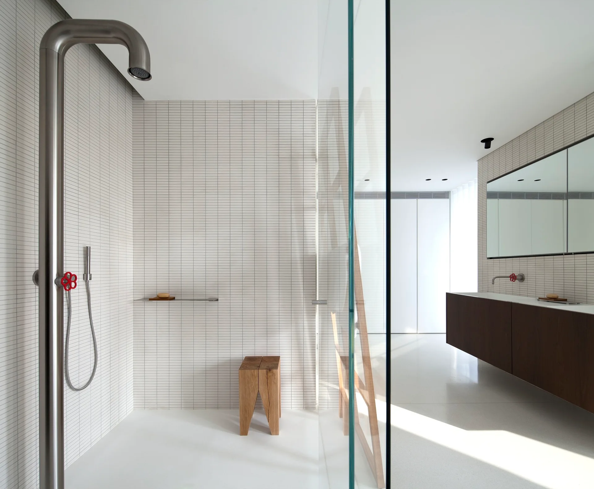 A minimalist walk-in shower features white grid tiles, a tall metal pipe showerhead with a red valve, and a small wooden stool.