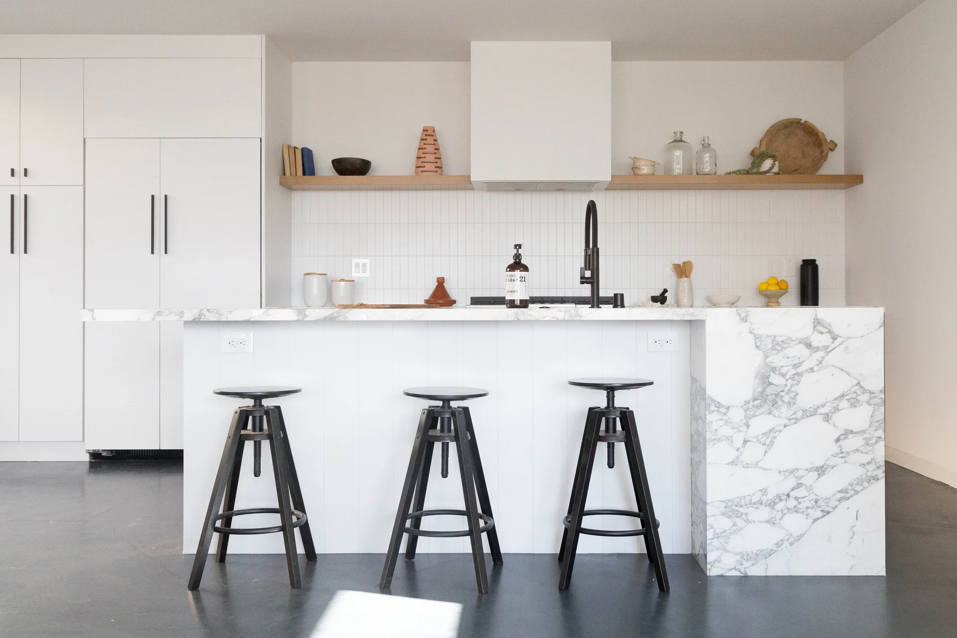 A minimalist kitchen features a white marble waterfall island with three black bar stools, a vertical tile backsplash, and a wooden floating shelf.