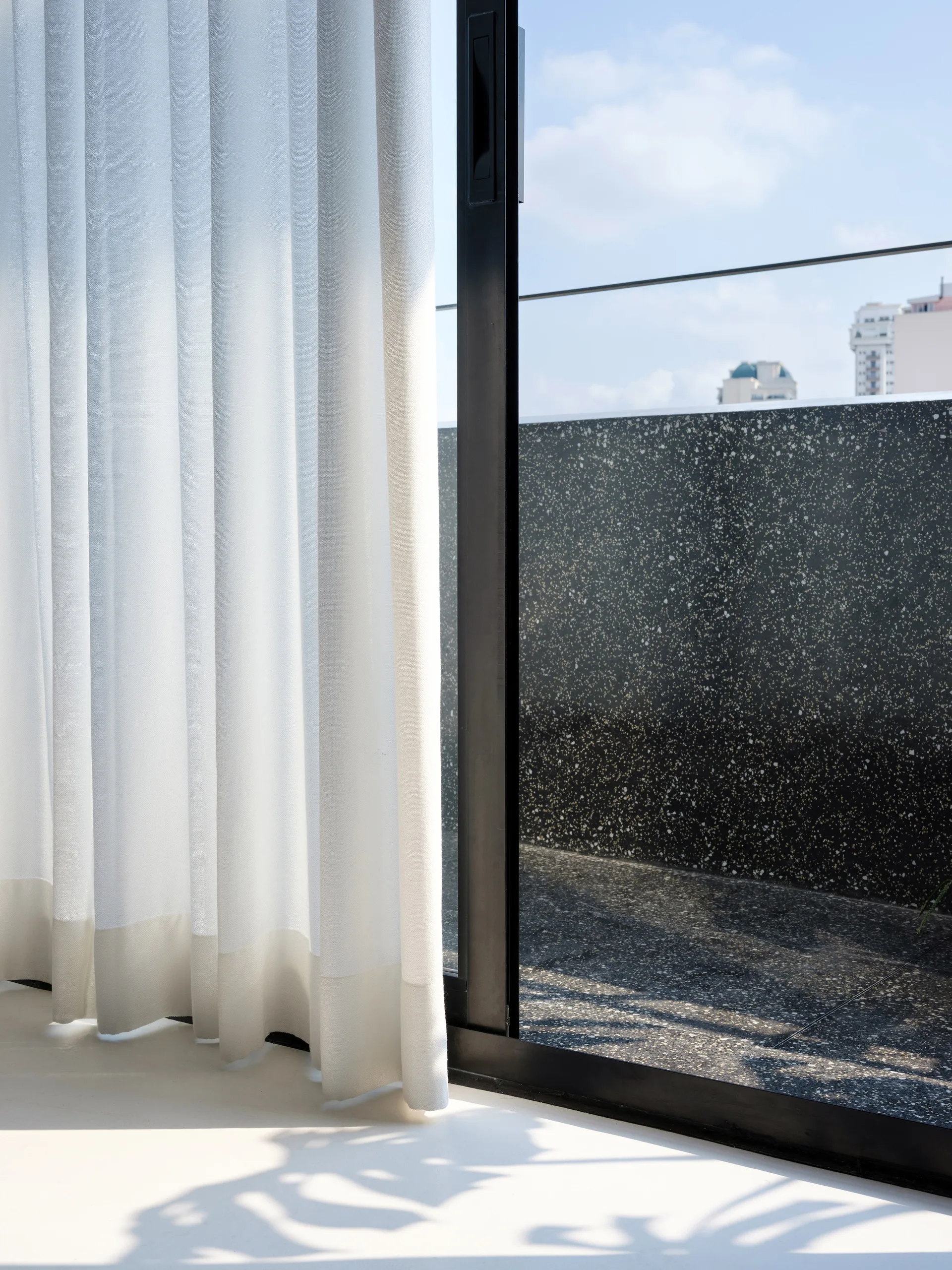 A black frame sliding glass door with a white curtain opens onto a terrace featuring dark speckled terrazzo flooring and a matching low wall.