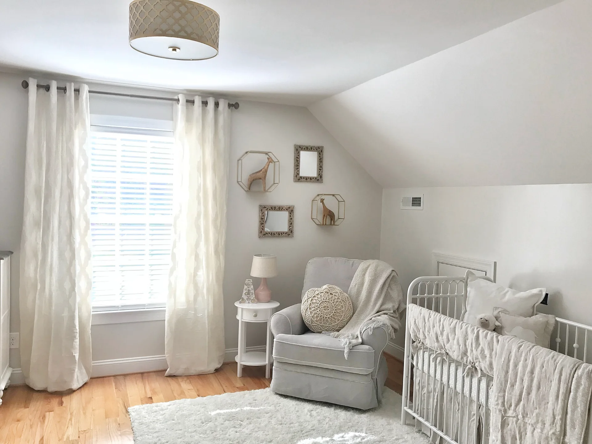 A light-filled attic nursery features a white metal crib, a grey glider armchair, and geometric wall shelves beneath a sloped ceiling.