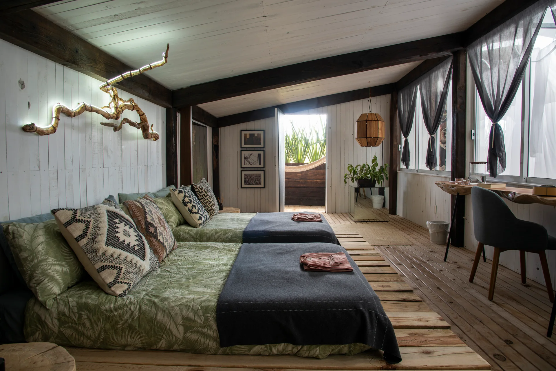Two low beds on wooden pallets sit in a rustic bedroom featuring white-paneled walls, dark exposed ceiling beams, and a live-edge desk by the window.