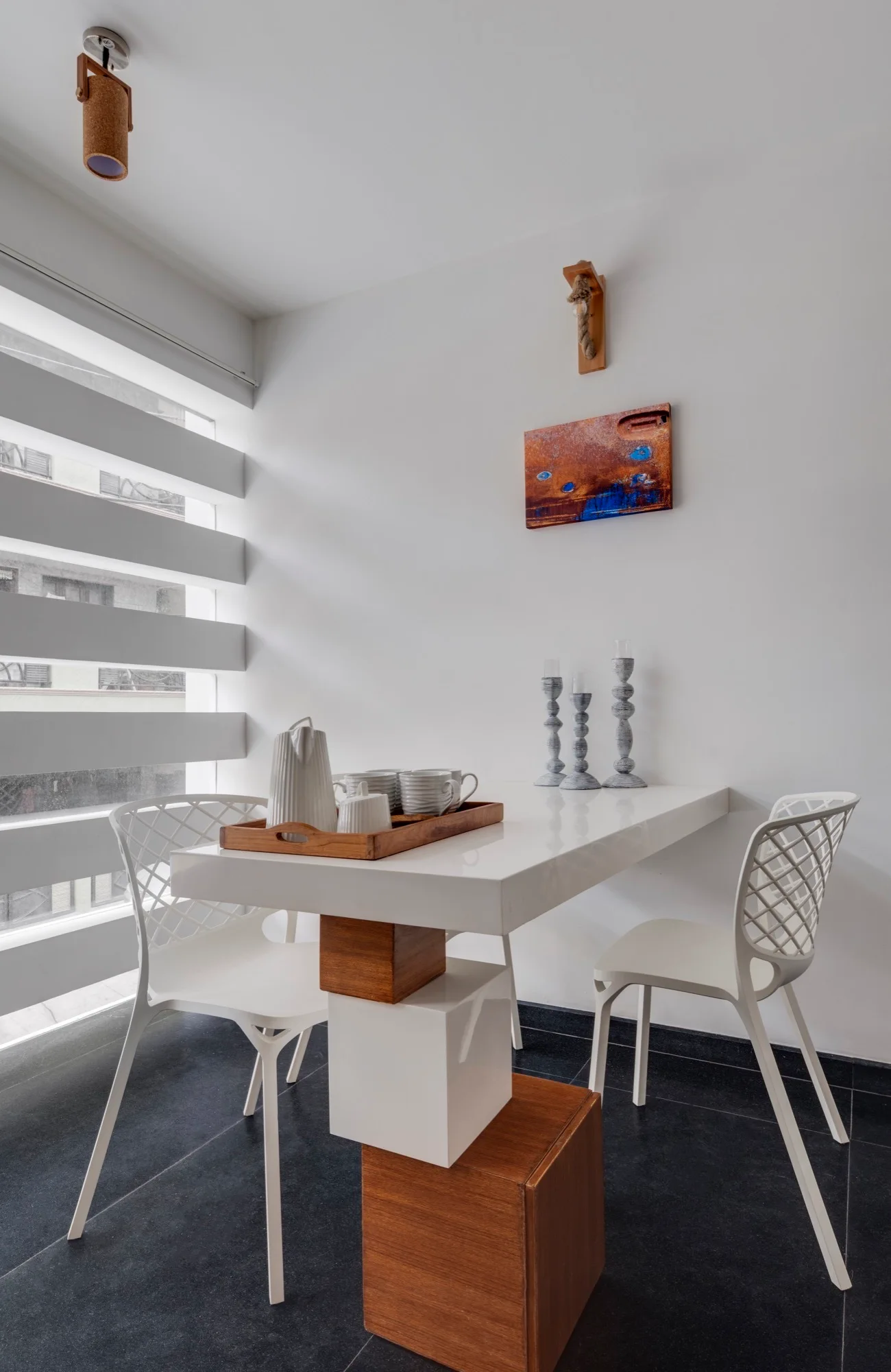 A white high-gloss dining table with a wood-accented block base is flanked by two white lattice-back chairs against a plain white wall.