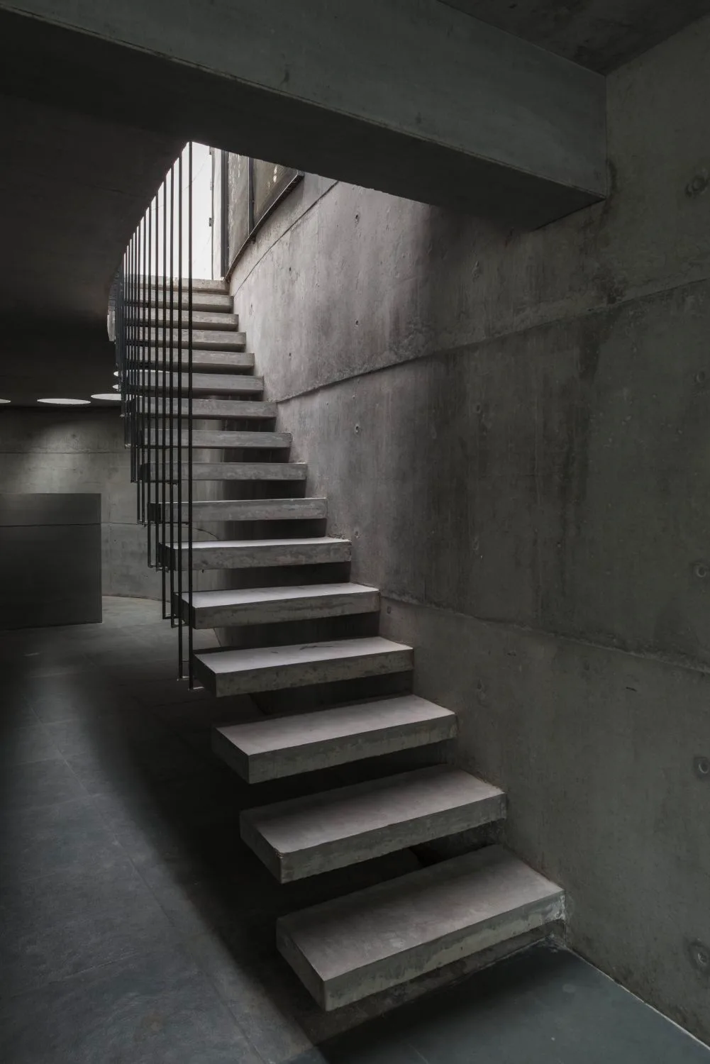 A modern cantilevered concrete staircase with thin vertical metal railings is set against a raw concrete wall.