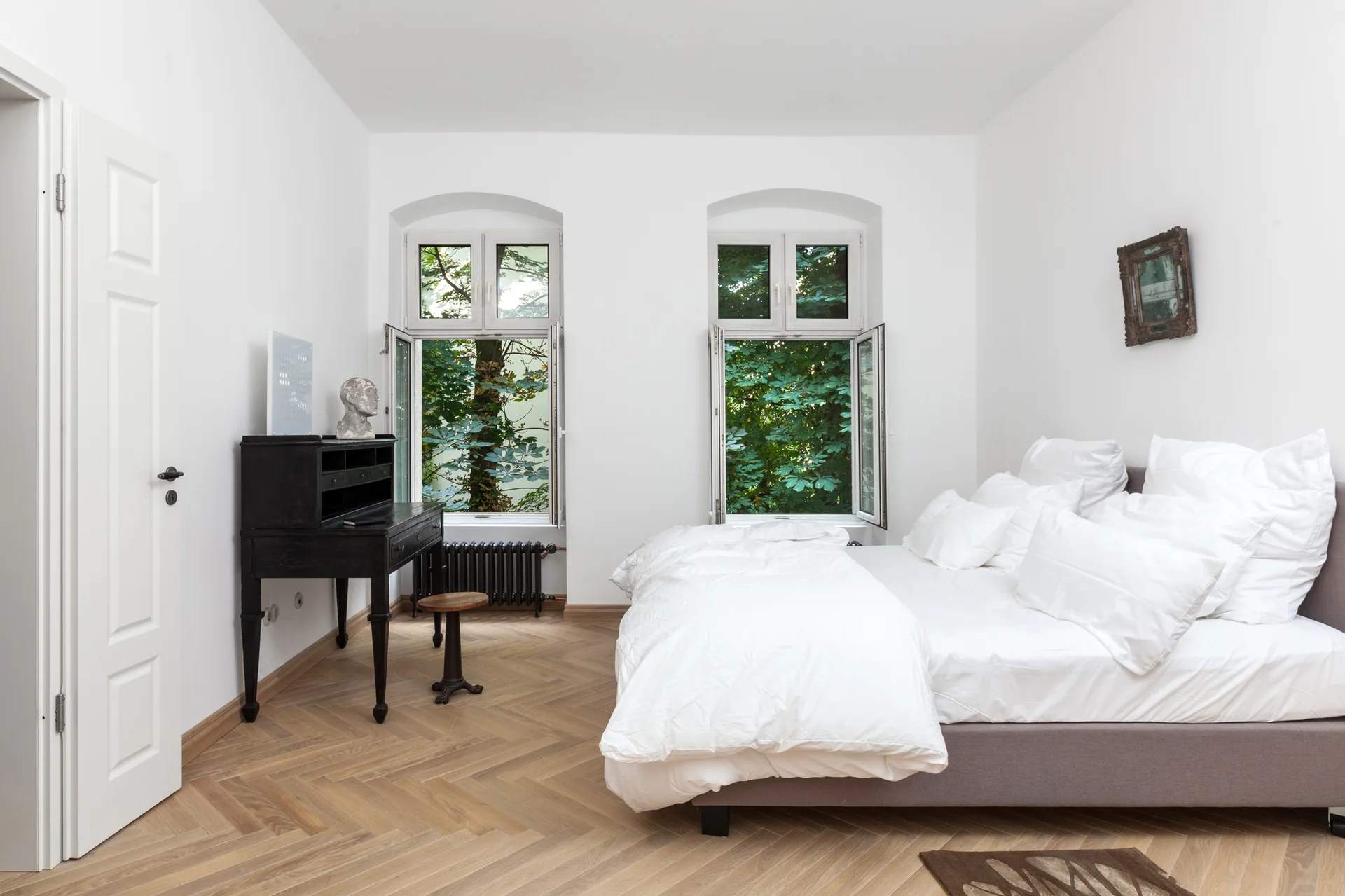 A bright bedroom featuring a bed with white linens, a black antique desk with a stool, and herringbone wood floors under two open windows.