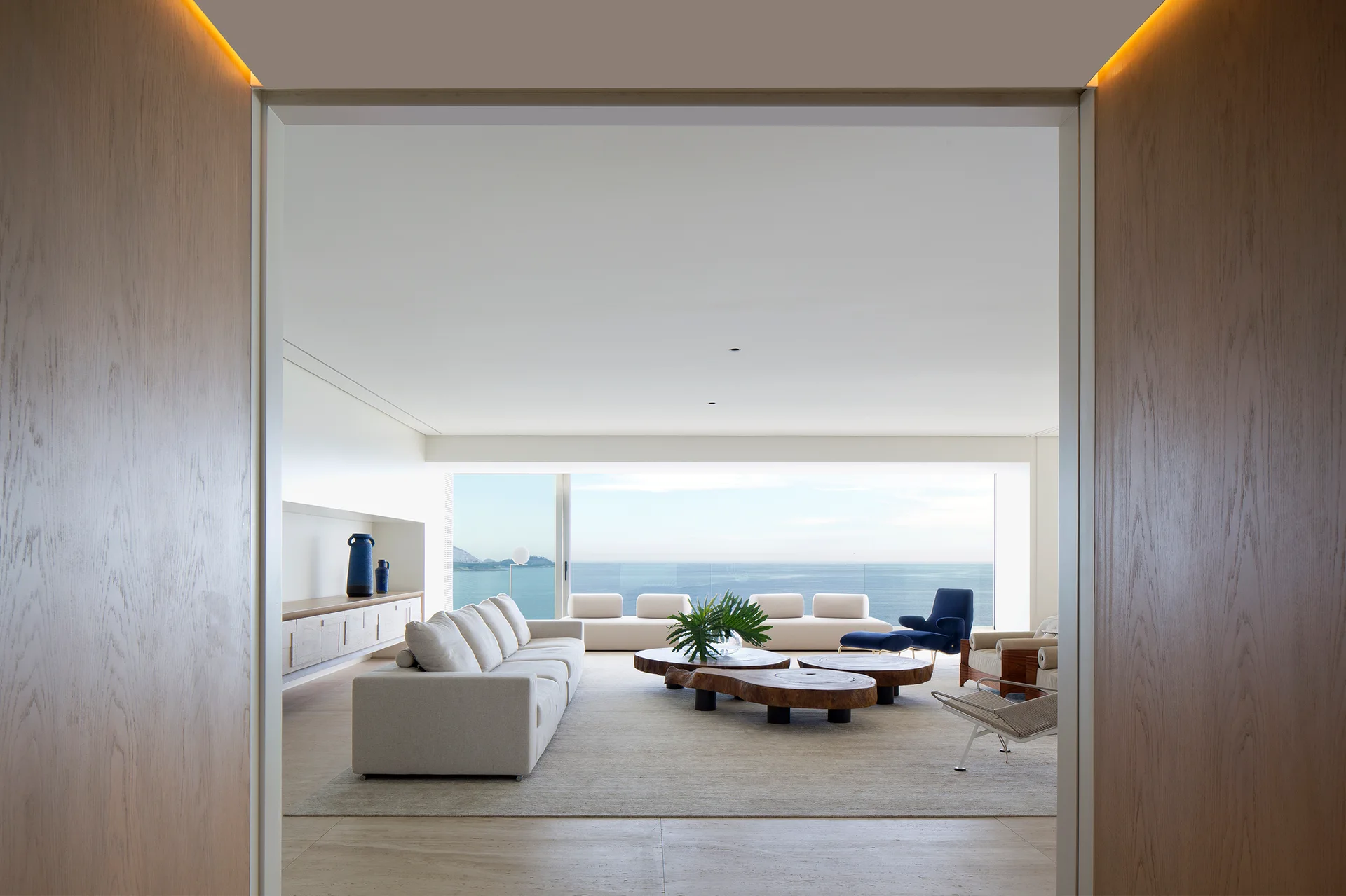 A modern living room with a cream sofa, live-edge wood coffee tables, and a large window looking out onto a blue ocean.