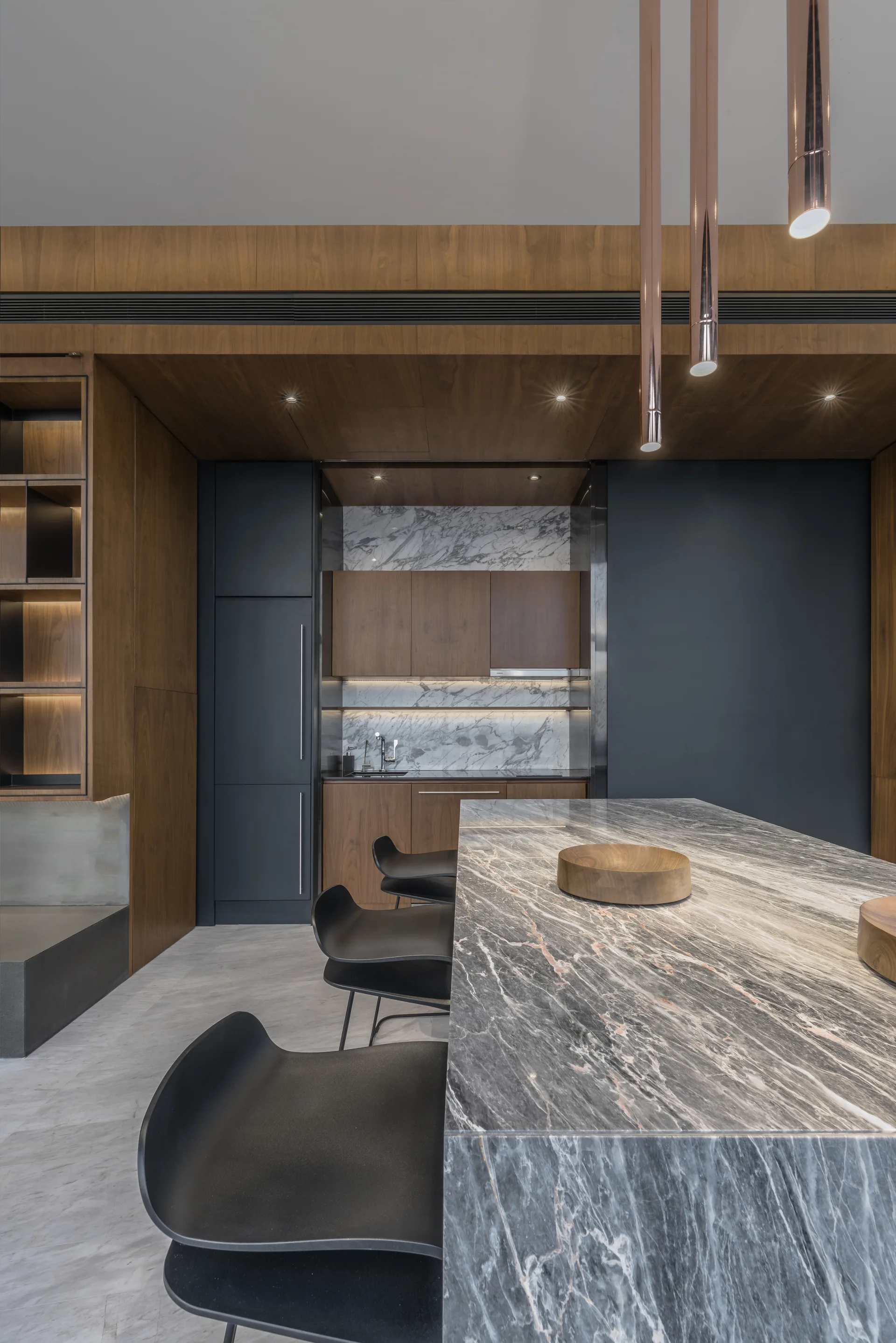 A modern kitchen features a grey veined marble island with black stools, copper pendant lights, and dark wood cabinetry with a marble backsplash.