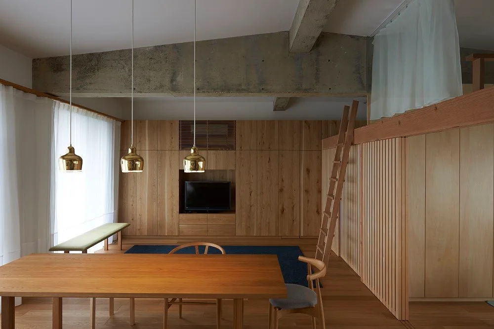 A modern wood-paneled living and dining area featuring a wooden dining table, three gold pendant lights, and an exposed raw concrete ceiling beam.