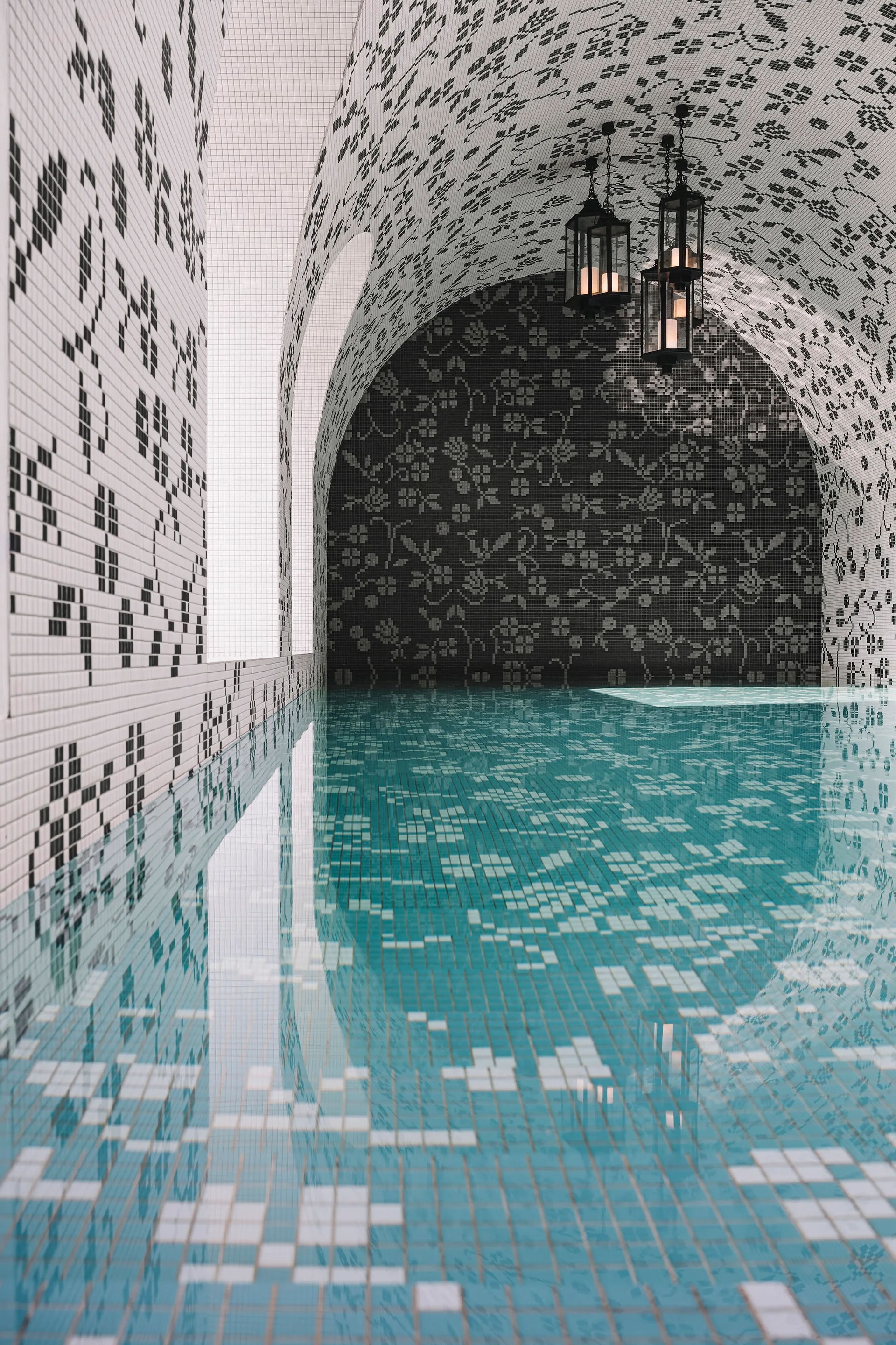 An indoor swimming pool features turquoise tiled water beneath a vaulted ceiling covered in intricate black and white floral mosaic tiles.