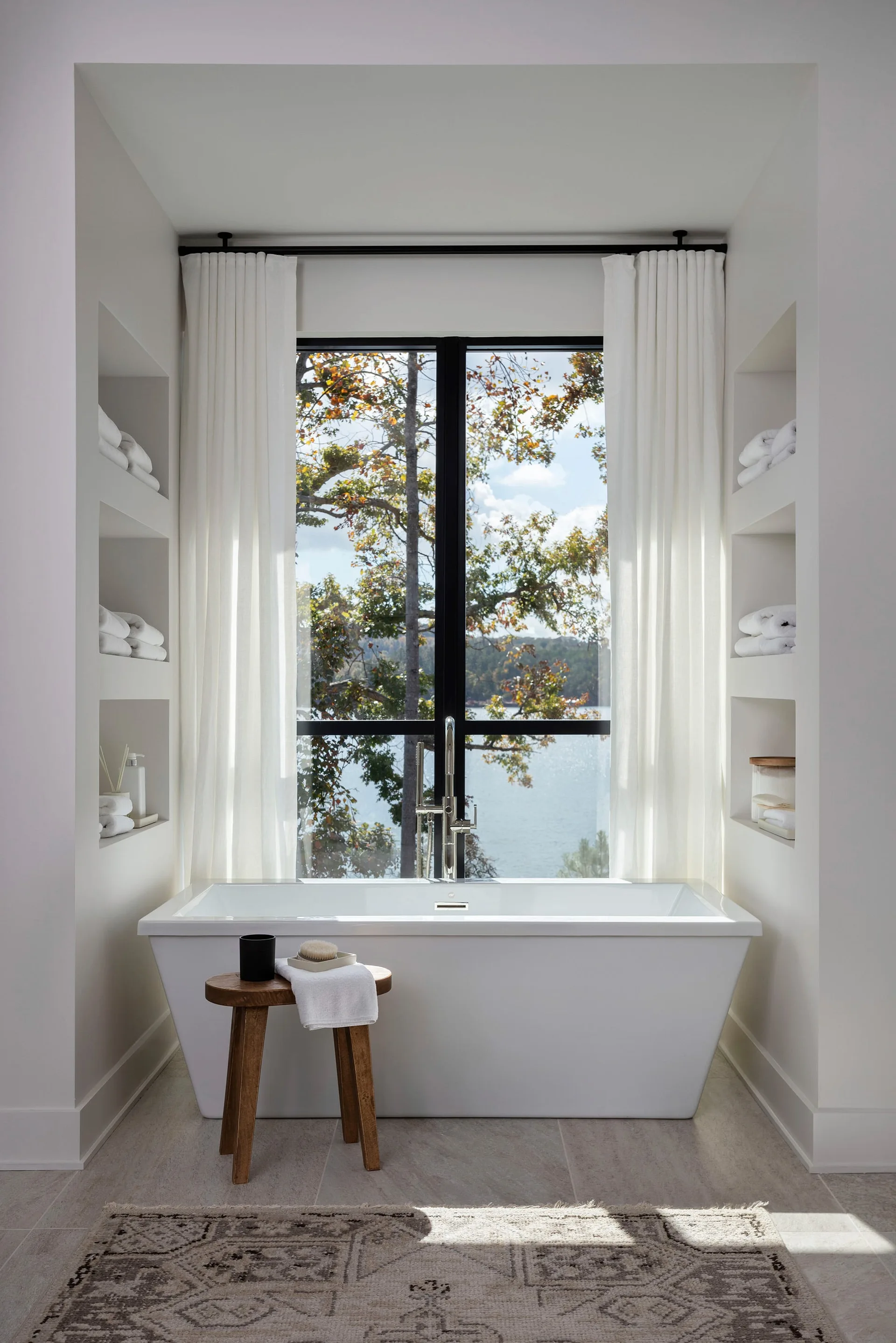 A white freestanding bathtub sits before a large window with water views, flanked by built-in wall niches filled with folded white towels.