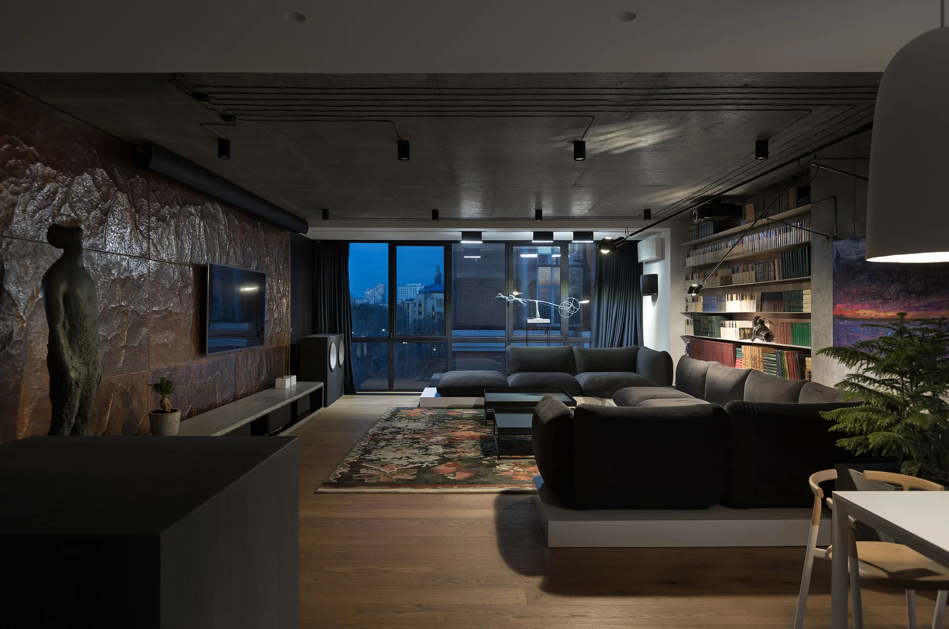 An industrial style living room featuring a dark grey sectional sofa, a textured metal wall with a television, and a concrete ceiling.