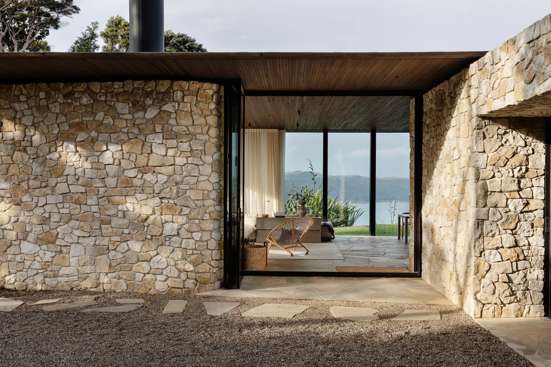 A stone patio with a gravel border leads into a living room flanked by rough-cut stone walls under a wood-slatted ceiling.