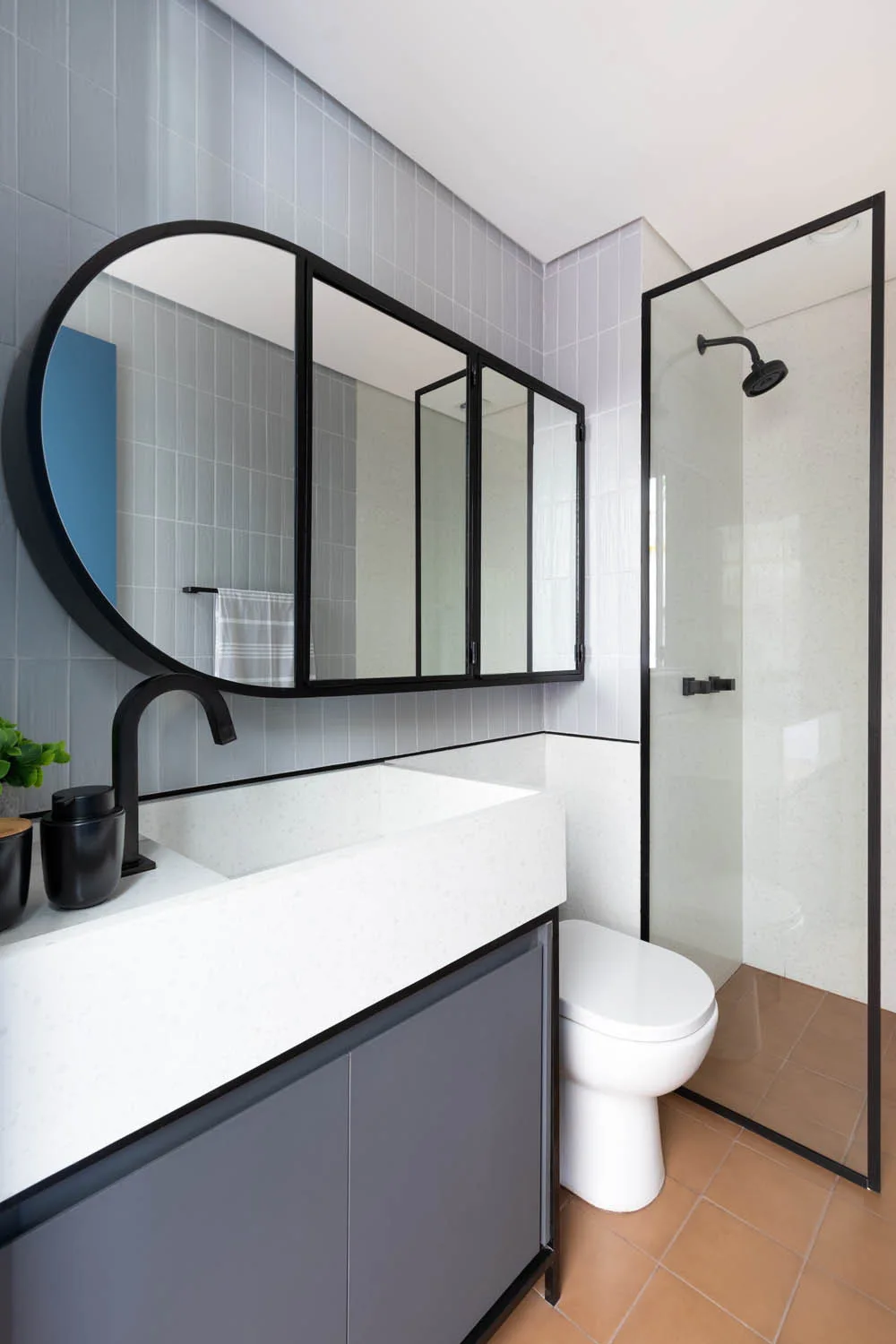 A modern bathroom featuring a white vanity with a black faucet, a large black-framed arched mirror, vertical grey wall tiles, and a glass shower.