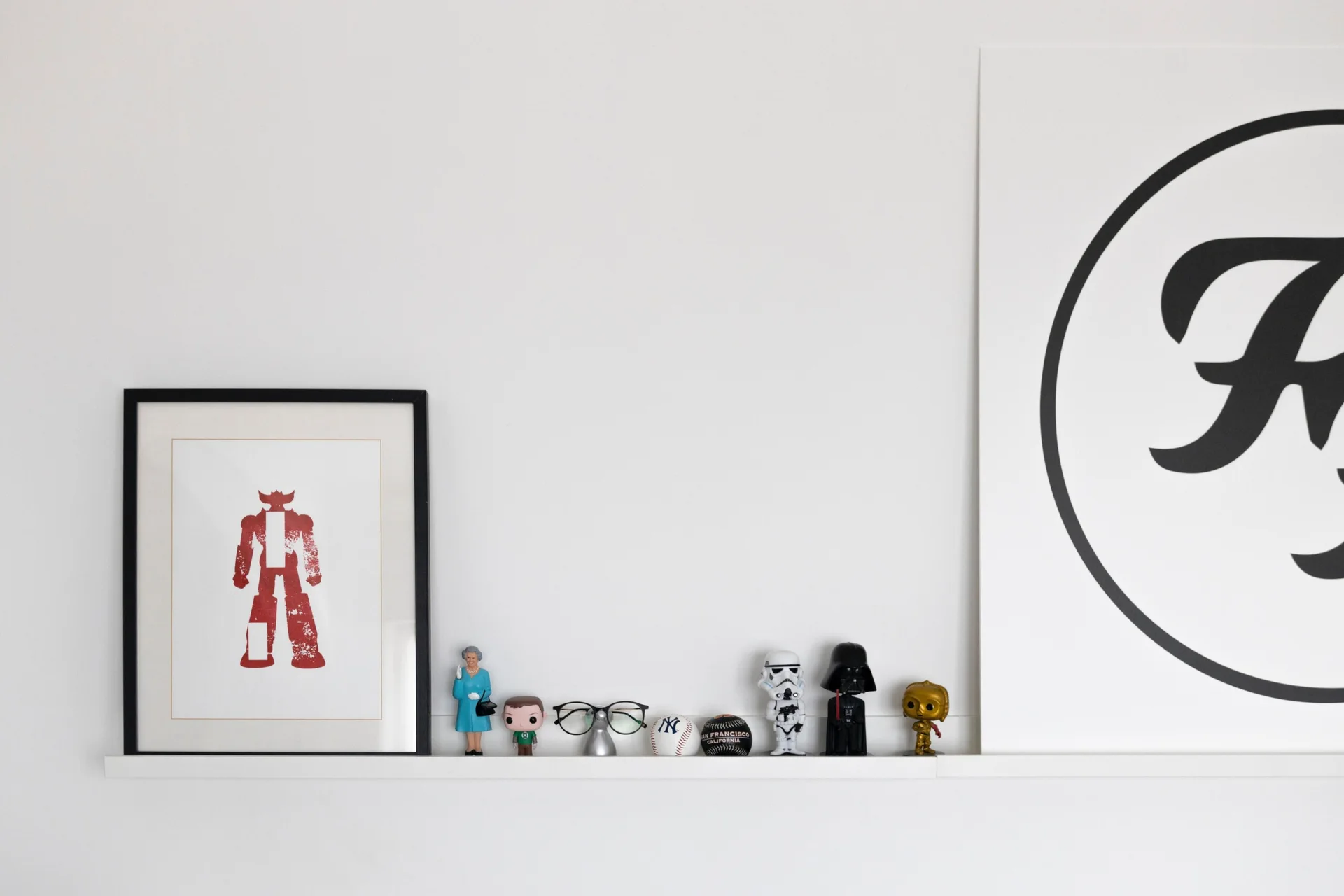 A white floating shelf mounted on a white wall holds framed graphic art, baseballs, and various small pop culture figurines.