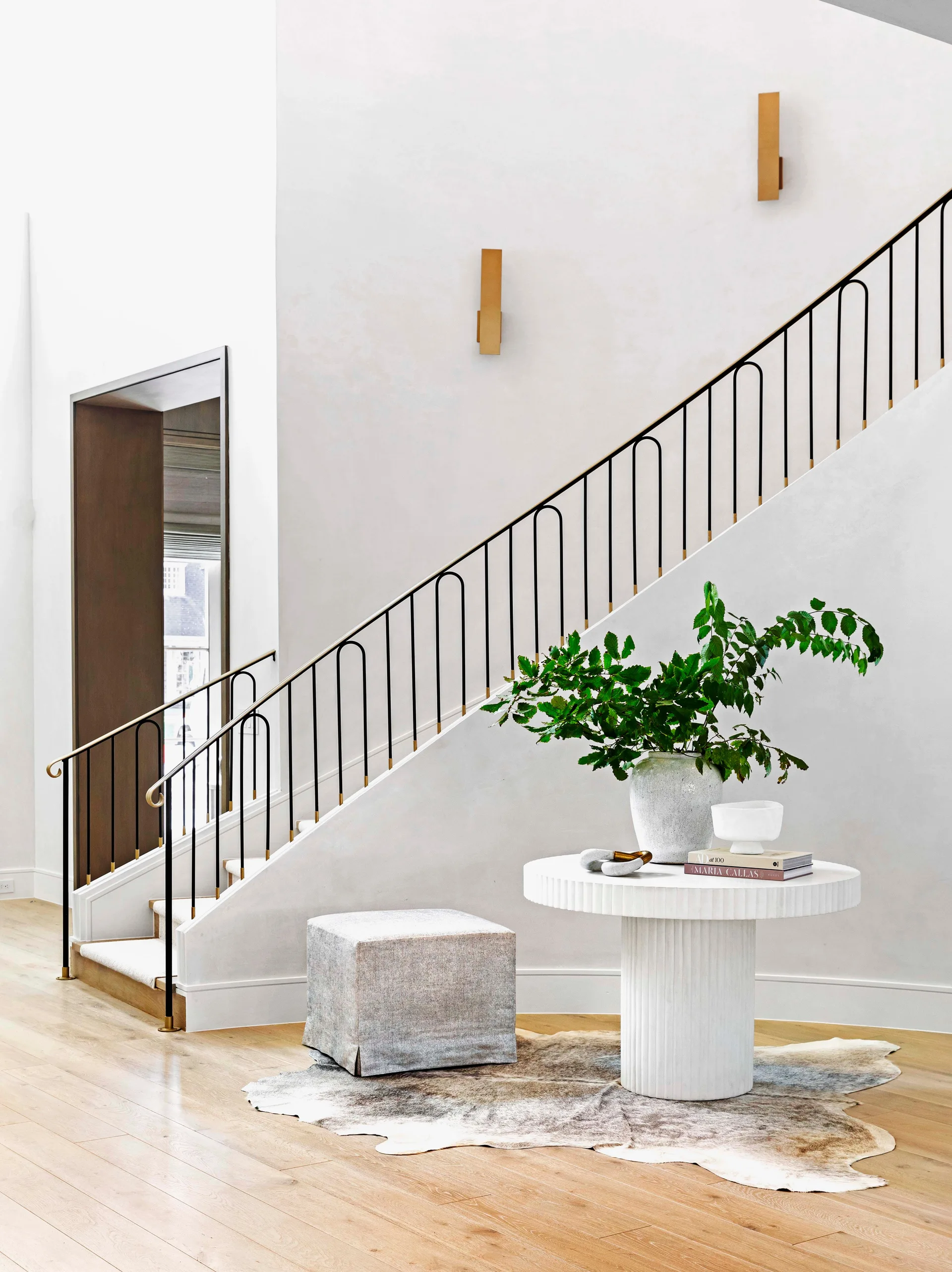 A modern entryway features a white fluted pedestal table on a cowhide rug next to a staircase with a black arched iron railing.