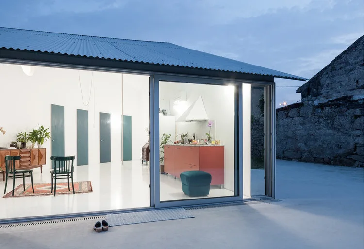 A modern open-plan kitchen and living space with glossy white floors, red cabinetry, and green accent doors viewed through a large sliding glass door.