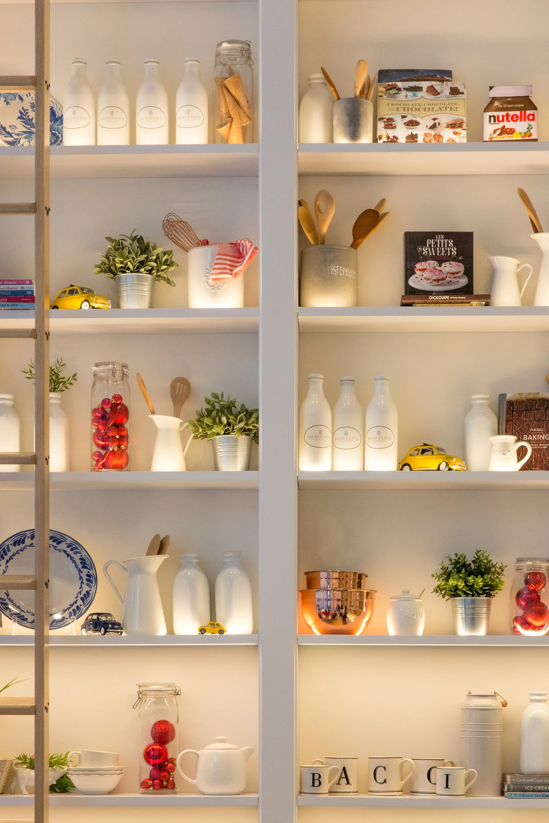 A tall white built-in kitchen shelving unit with integrated LED lighting displaying organized glassware, cookbooks, potted plants, and ceramic pitchers.