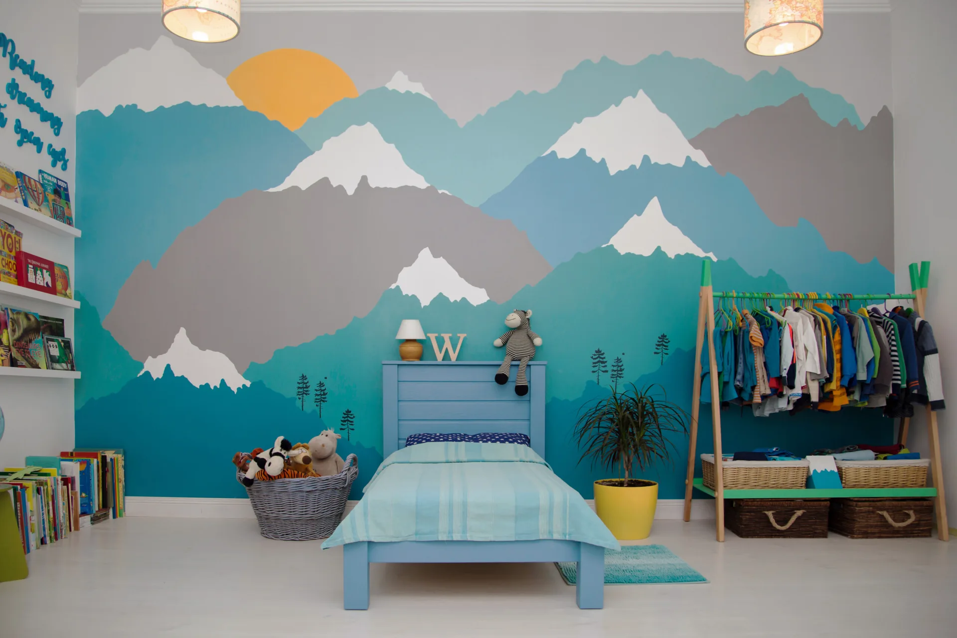 A kids bedroom featuring a blue wooden bed set against a colorful mountain mural with an adjacent light wood clothing rack.