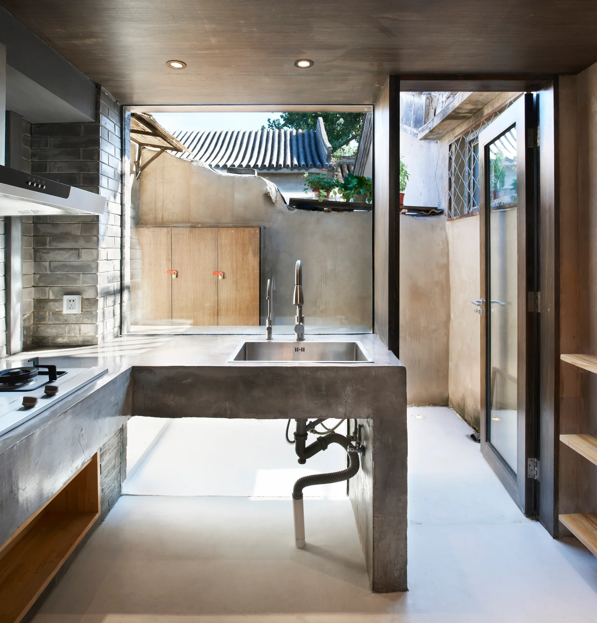 A minimalist industrial kitchen featuring a cast concrete countertop with a sink, exposed gray brick walls, and a large window overlooking a courtyard.