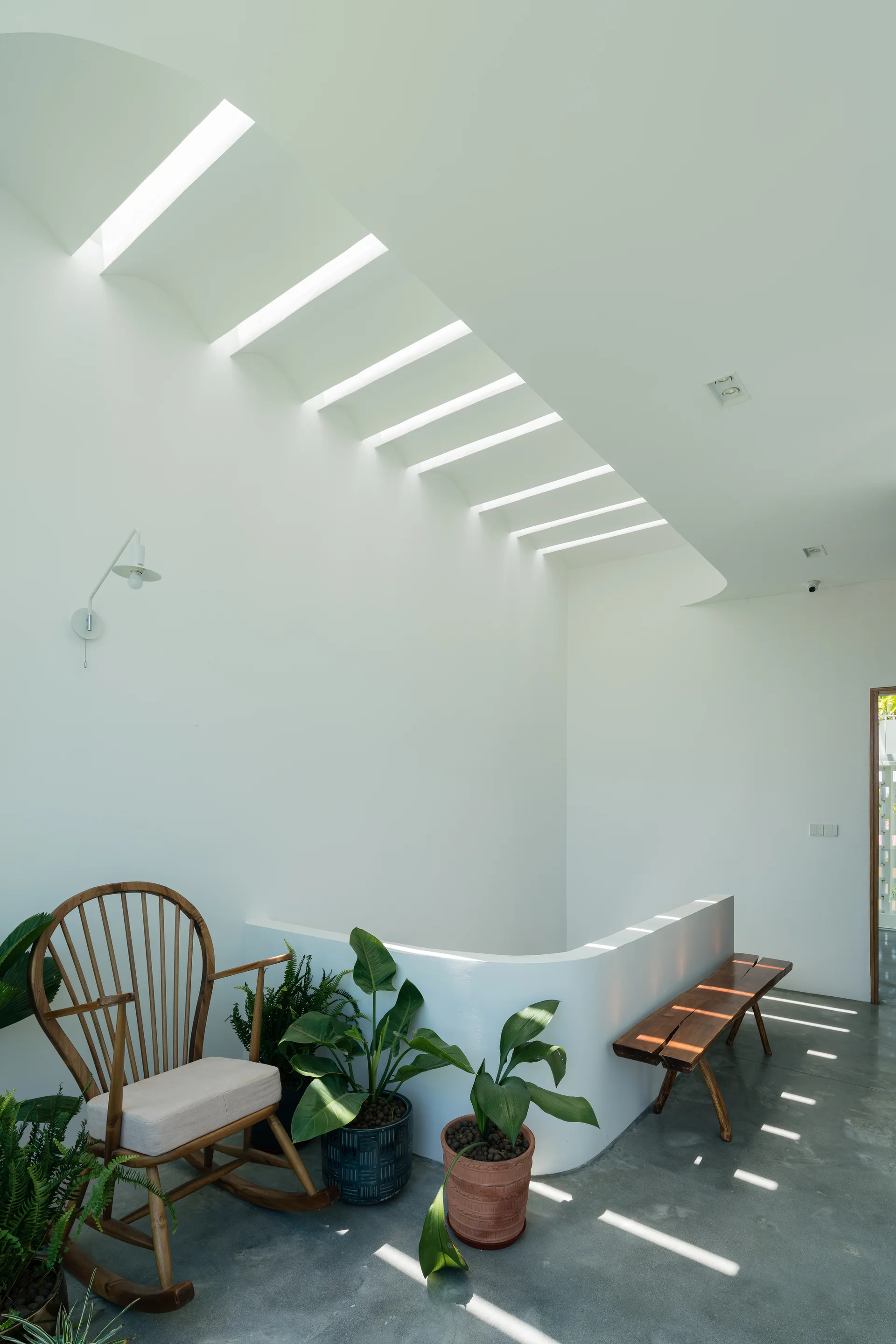 A minimalist white stair landing features a wooden rocking chair, a wooden bench, potted plants, and light filtering through a slatted skylight.