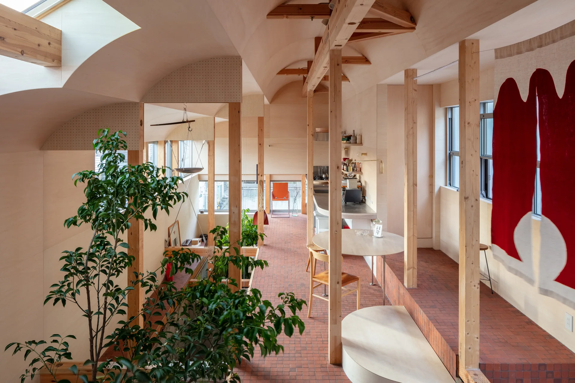 An airy interior space featuring an arched plywood ceiling, exposed timber columns, stepped terracotta tile floors, and natural light from a skylight.