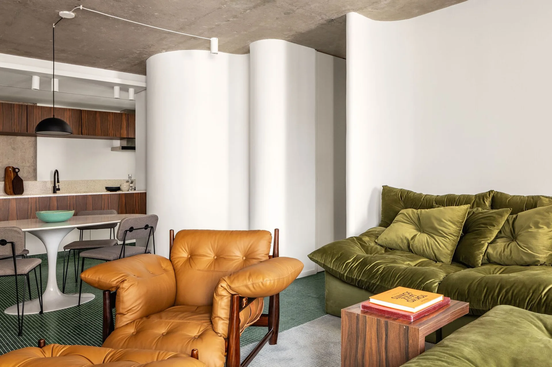 An open-plan living room featuring an olive green velvet sofa, a tan leather armchair, a curved white partition wall, and small green floor tiles.