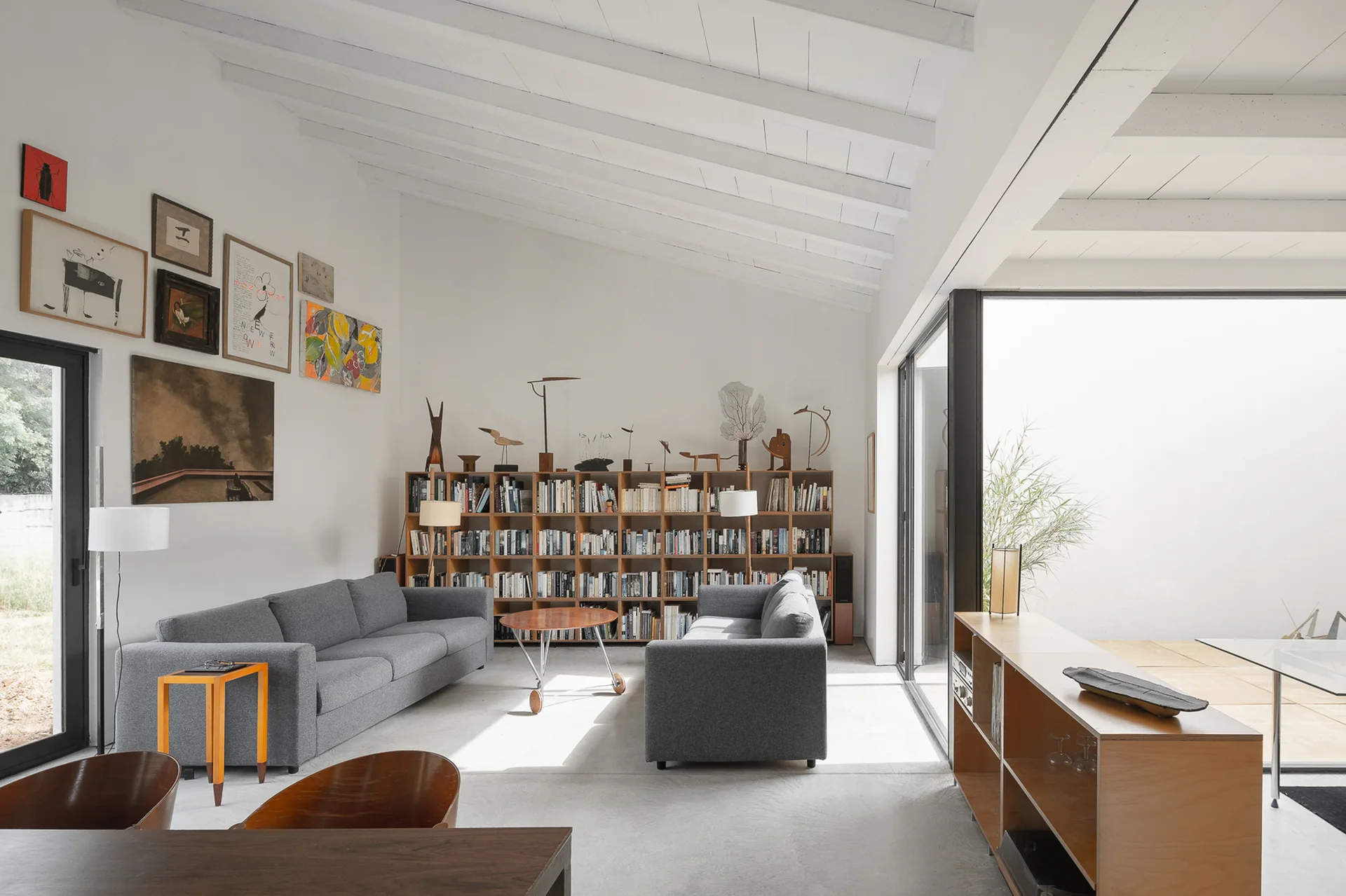 A modern living room features two gray sofas, a low wooden bookcase, and a sloped white ceiling with exposed wooden beams.