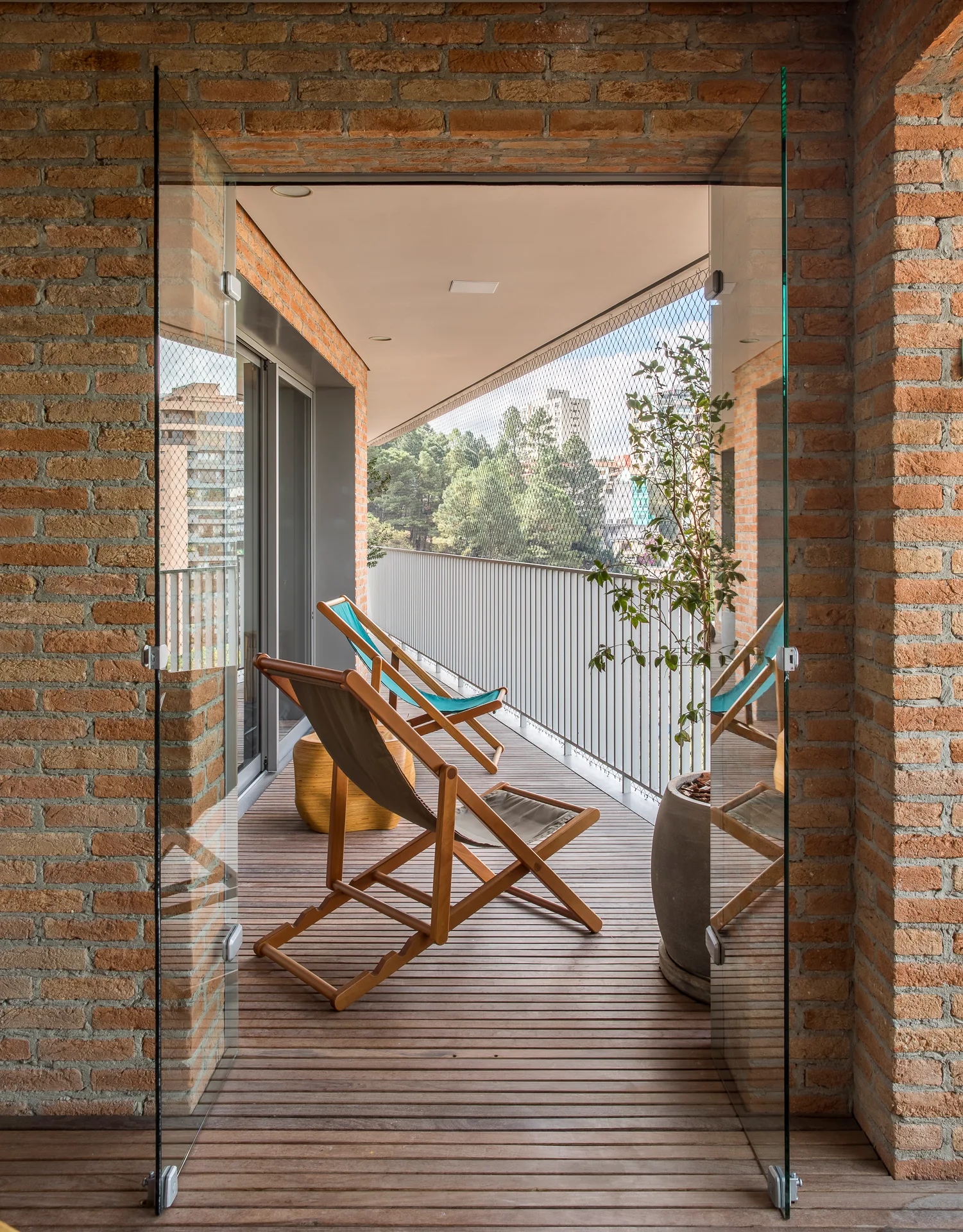 Frameless glass pivot doors open to a sunlit balcony featuring wood deck flooring, exposed brick walls, and two folding lounge chairs.