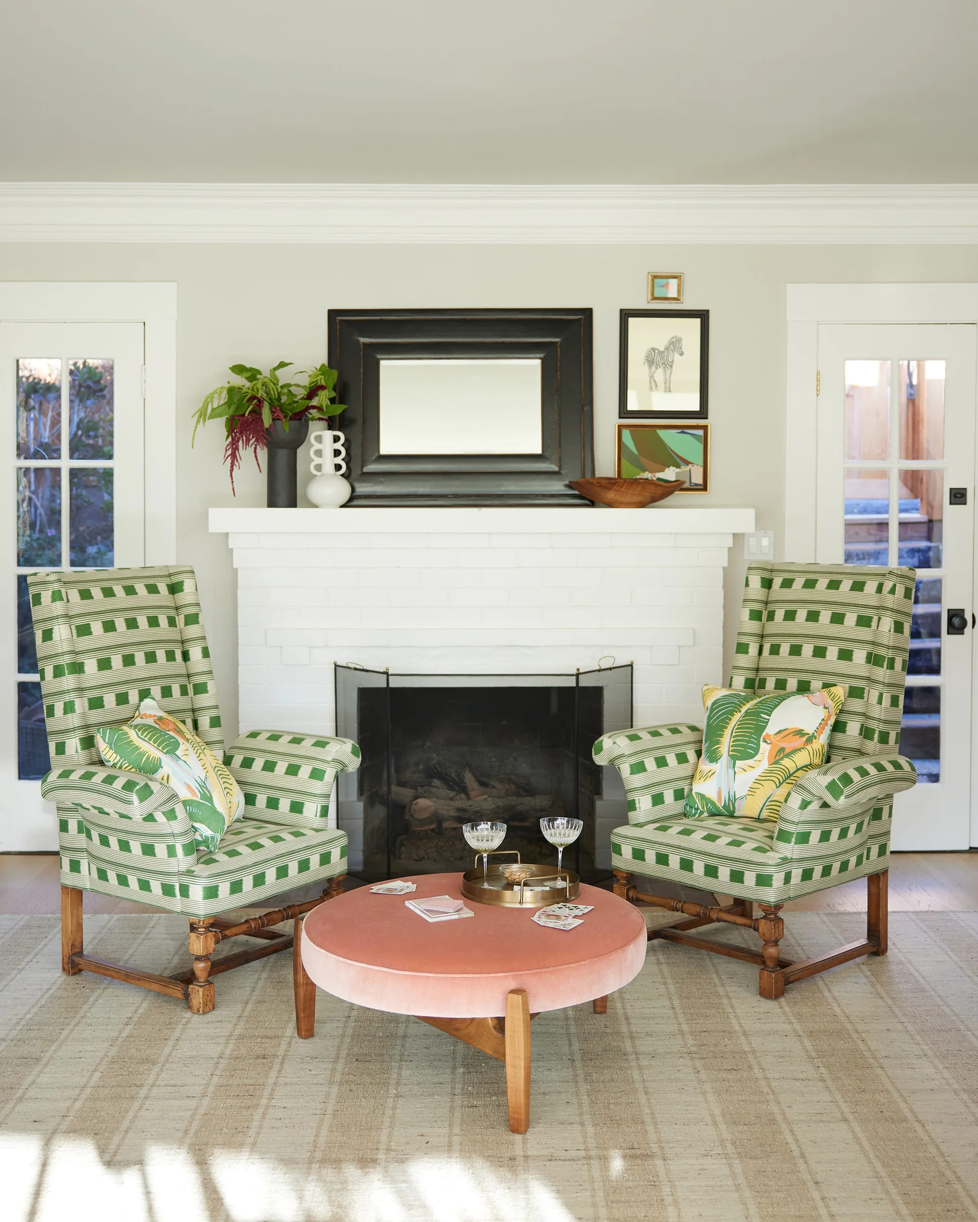 Two green checked armchairs with tropical print pillows sit next to a white brick fireplace, with a round pink velvet ottoman in the foreground.