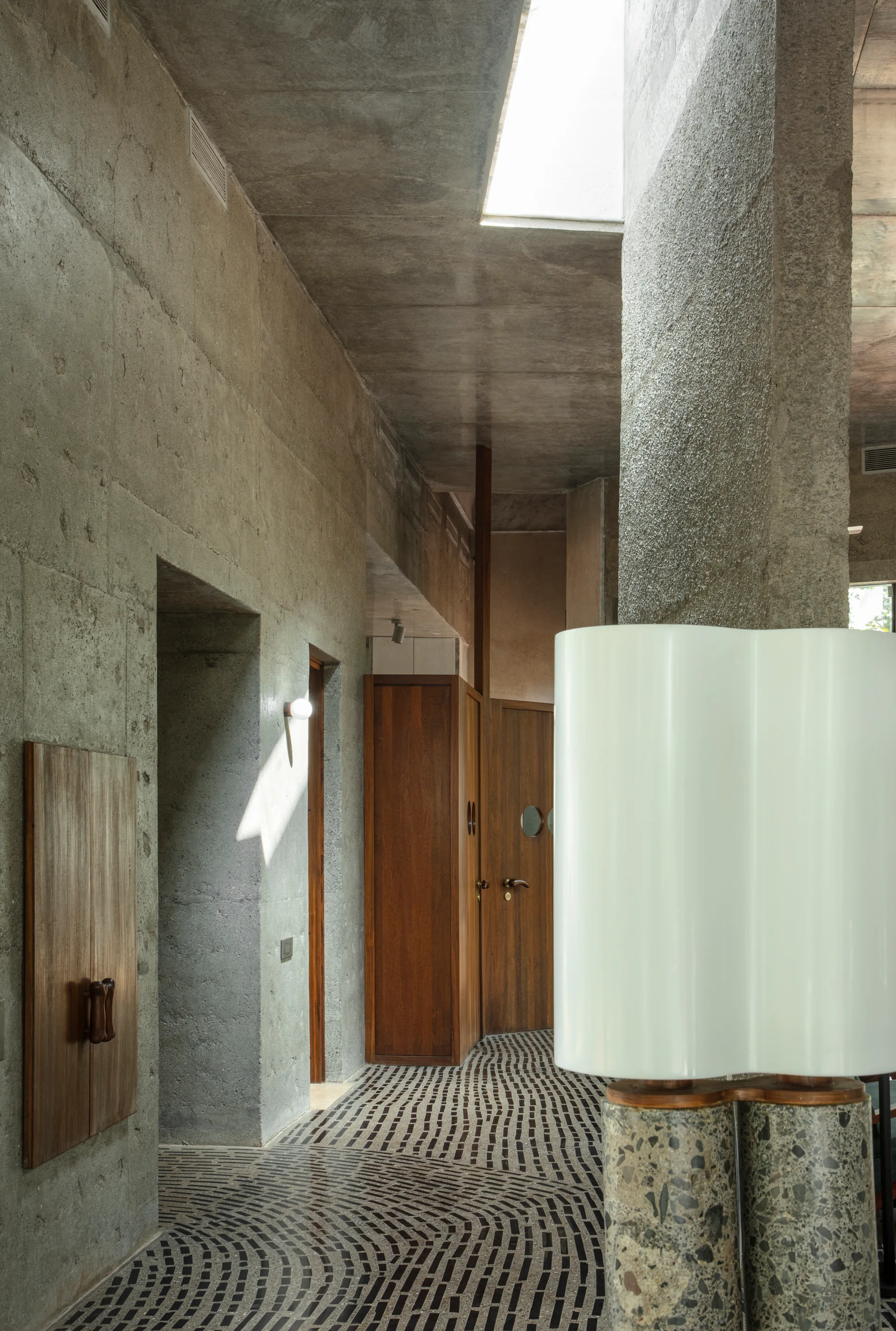 An interior hallway features raw exposed concrete walls, warm wood cabinets, a skylight, and a curved black and white mosaic tile floor.