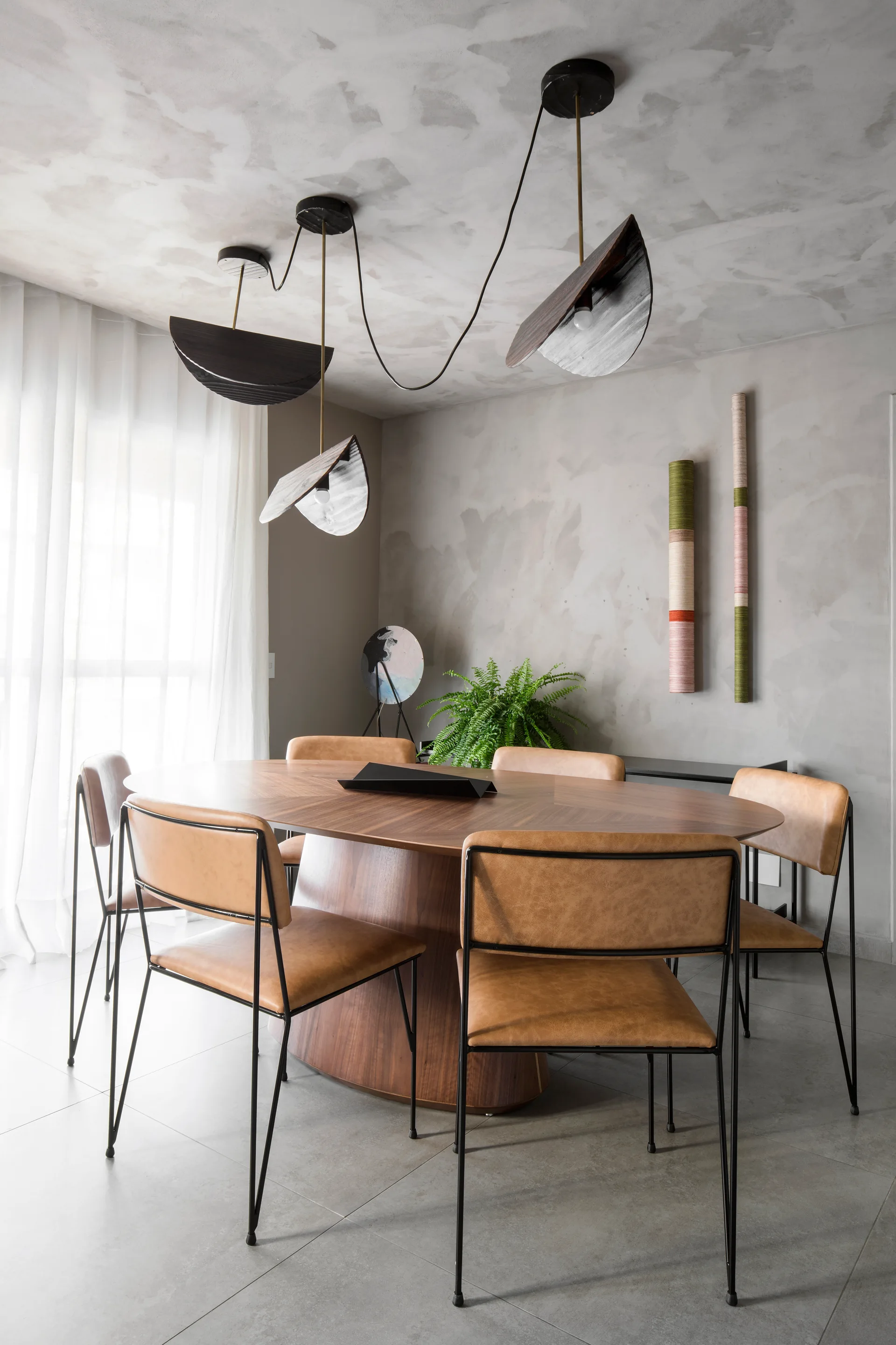 An oval wood dining table is surrounded by tan leather chairs under a sculptural pendant light in a room with textured gray plaster walls.