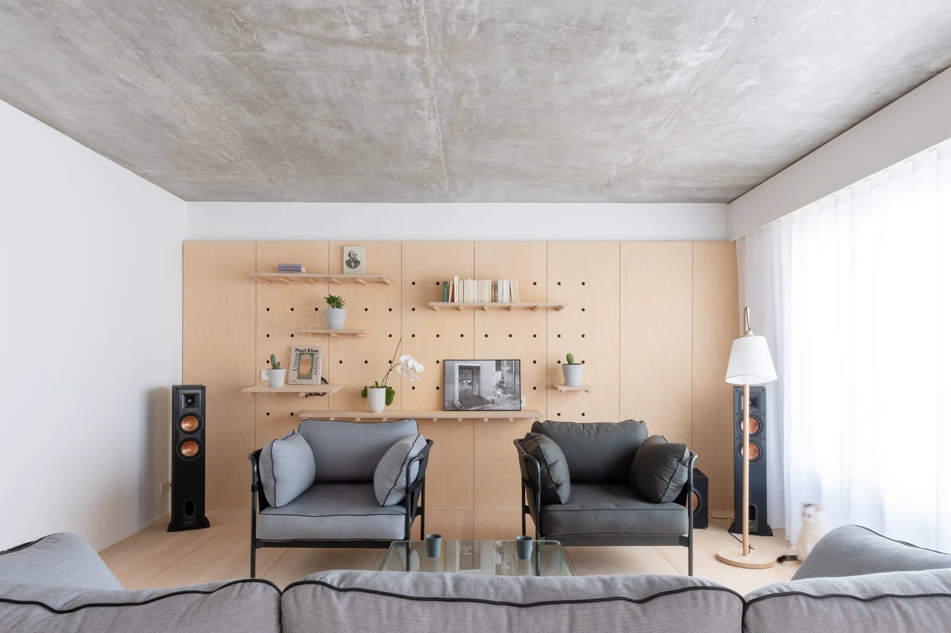 A modern living room features a large plywood pegboard wall with shelves, an exposed concrete ceiling, and two armchairs facing a glass coffee table.