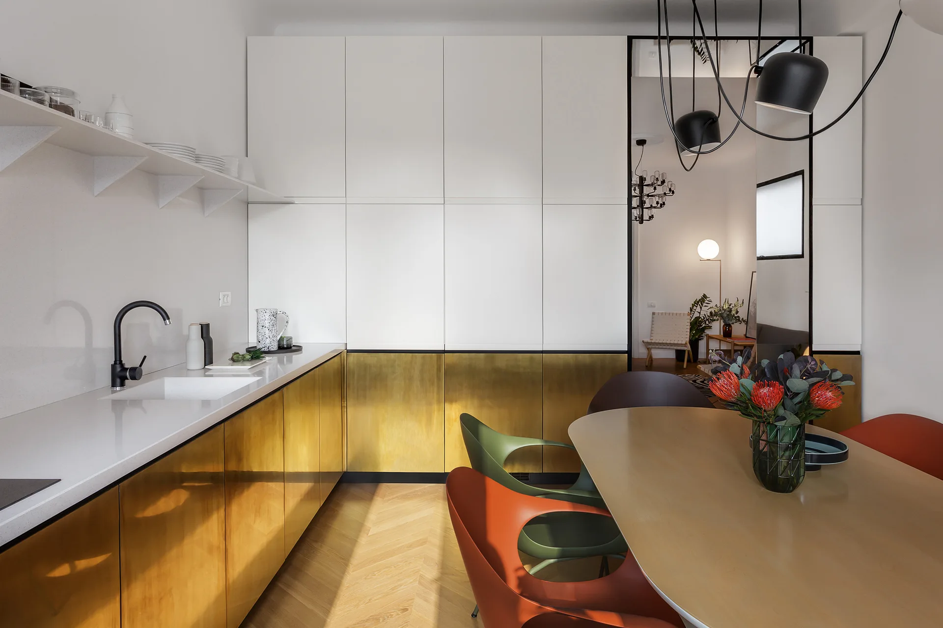 A modern kitchen and dining area features white and brass cabinetry, herringbone wood flooring, and a dining table with colorful cutout chairs.