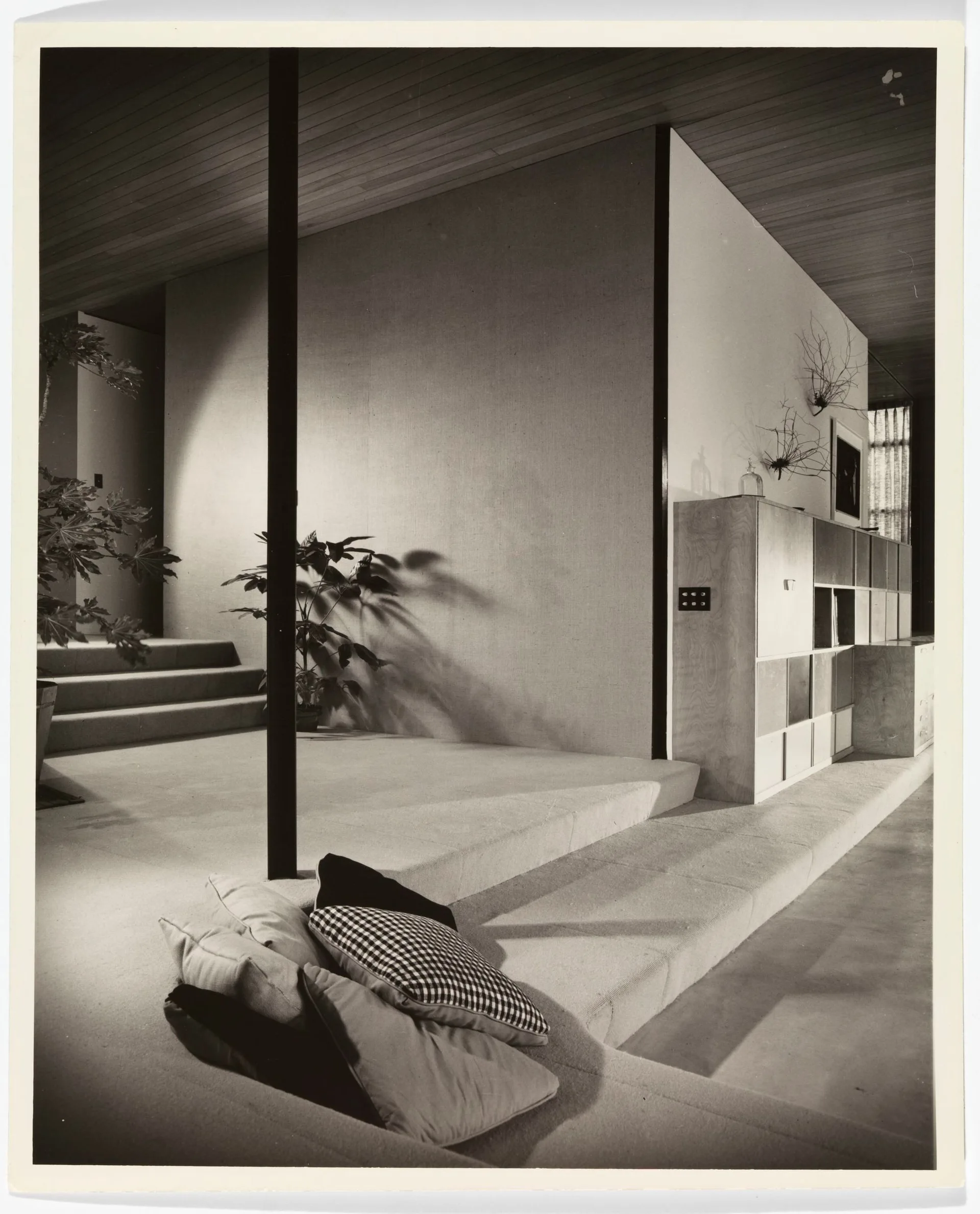 A black and white photograph of a mid-century sunken lounge with carpeted steps, a grid-style wooden storage cabinet, and a wood-planked ceiling.