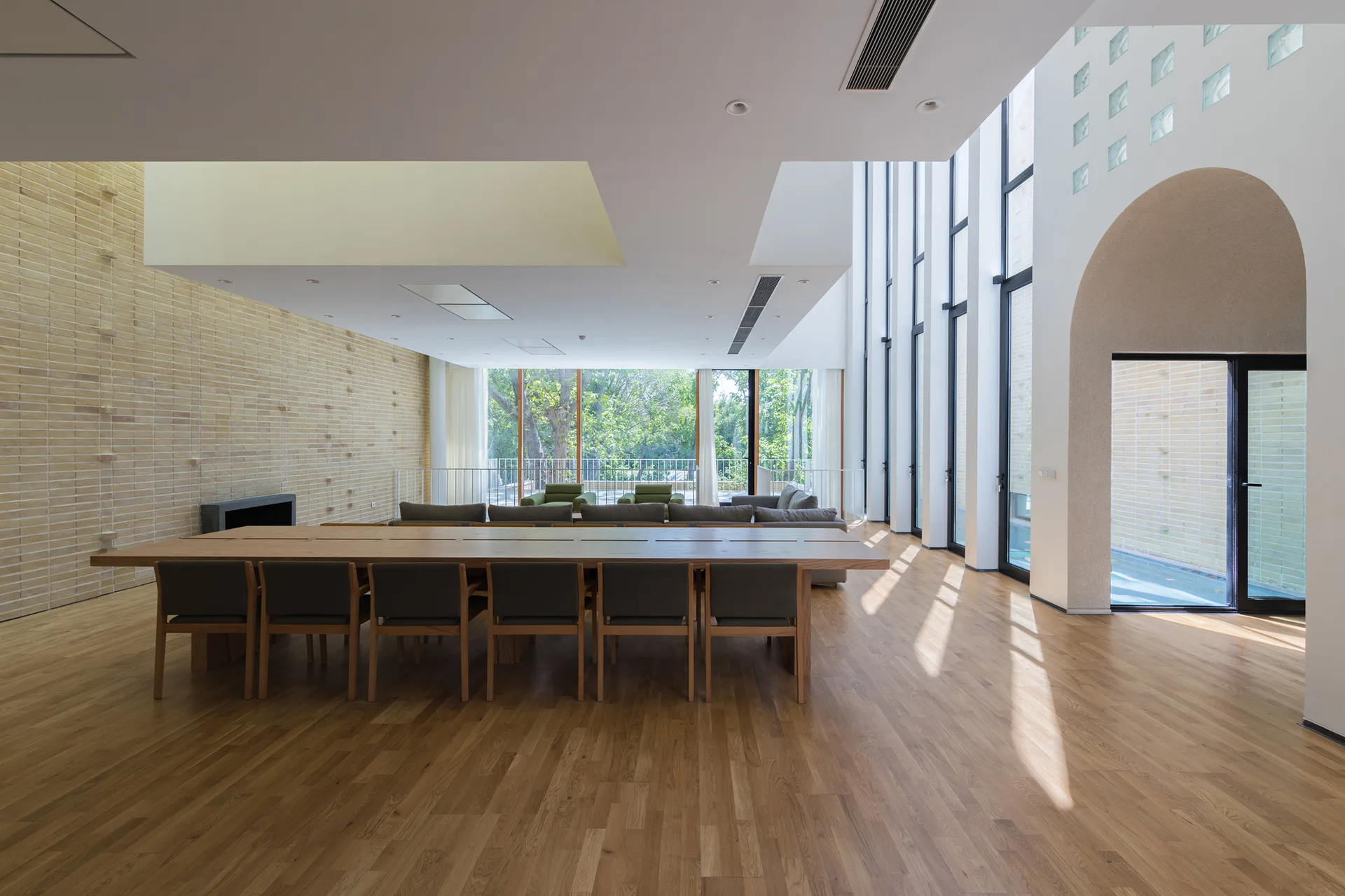 An open-plan room features a long wooden dining table, a textured brick wall with a fireplace, and a towering wall of windows.