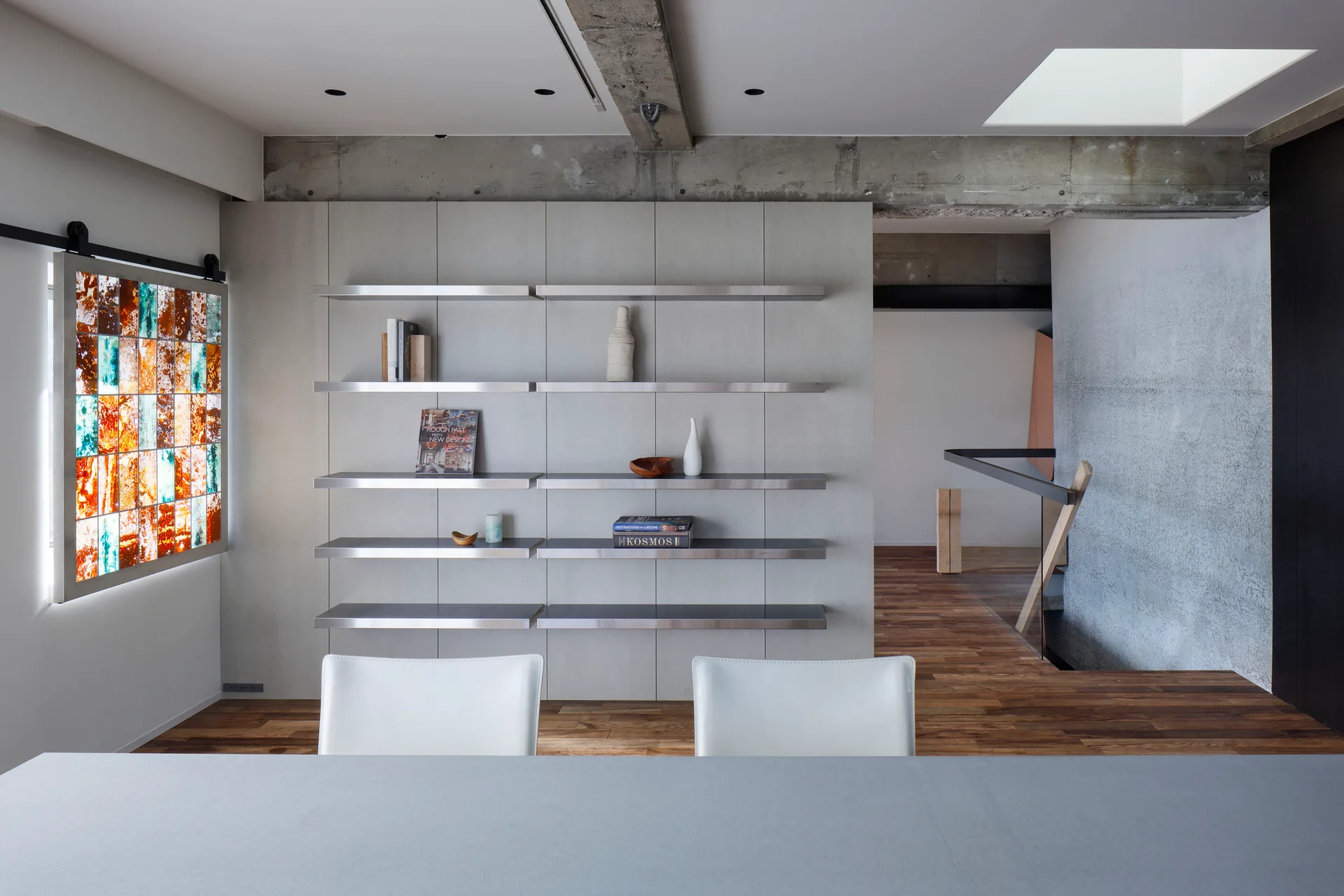 A minimalist dining room featuring a light gray paneled wall with metal shelves, an exposed concrete beam, and a colorful sliding stained glass panel.