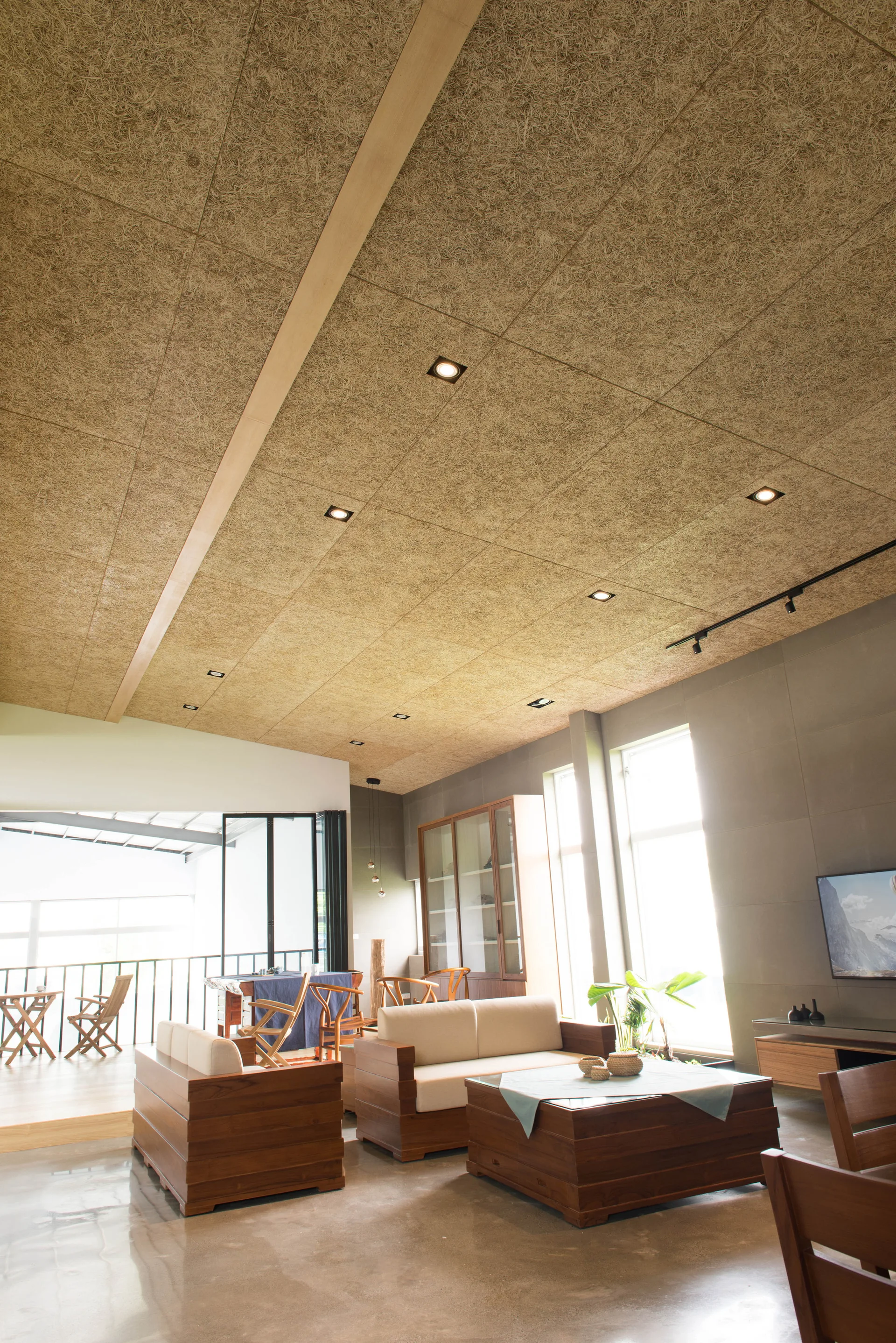 A modern living room features low wood-framed sofas, a polished concrete floor, and a textured wood-fiber ceiling with recessed lights.