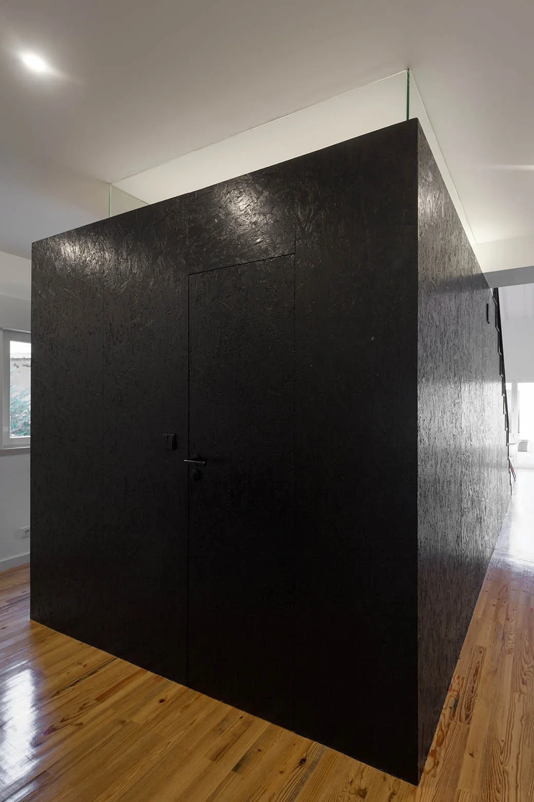 A central black OSB partition block with a flush door stands on glossy light wood flooring in a minimalist hallway.