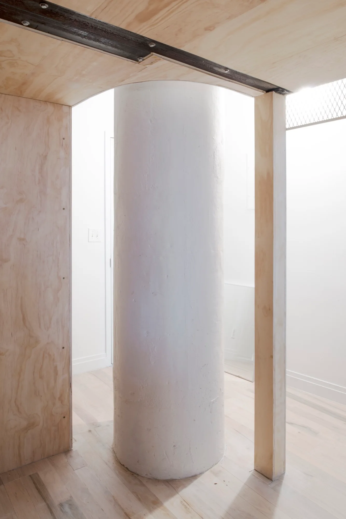 A white cylindrical support column is framed by raw plywood partition walls and an arched ceiling transition over light hardwood floors.