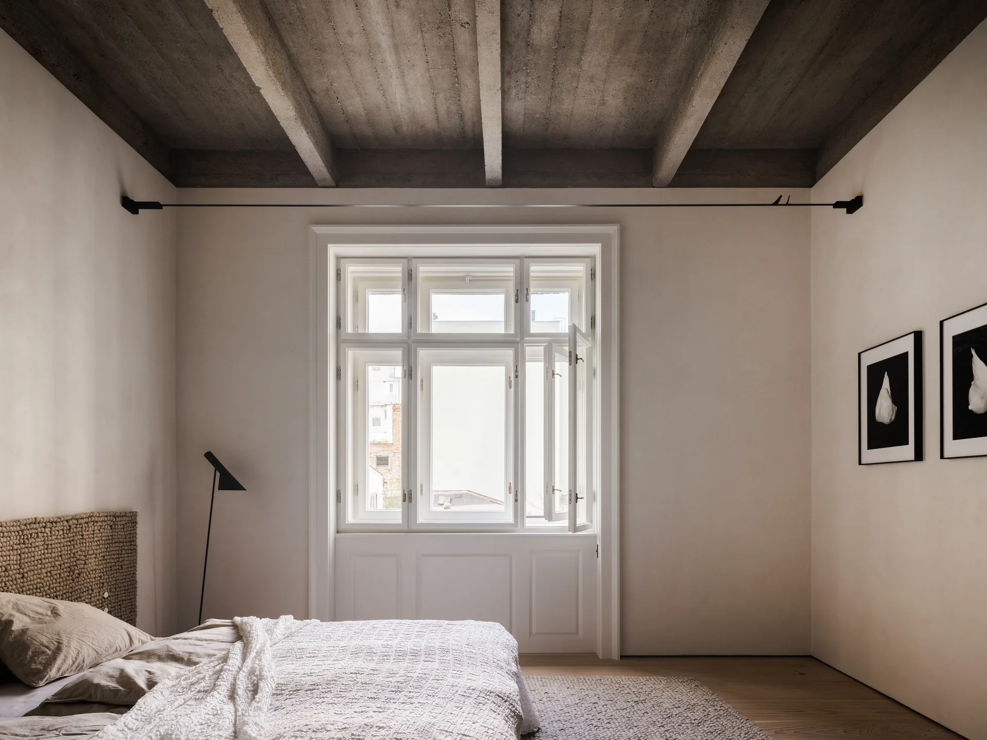 A minimalist bedroom featuring raw concrete ceiling beams, a textured woven headboard, and a large multi-pane window with white wood detailing.