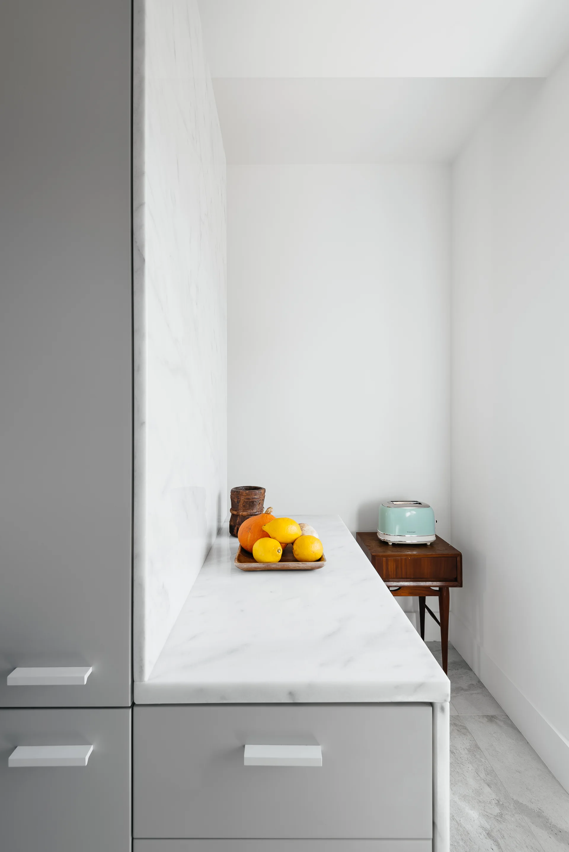 A minimalist kitchen detail featuring light grey cabinetry, a white veined marble countertop, a slab backsplash, and a small wooden side table.