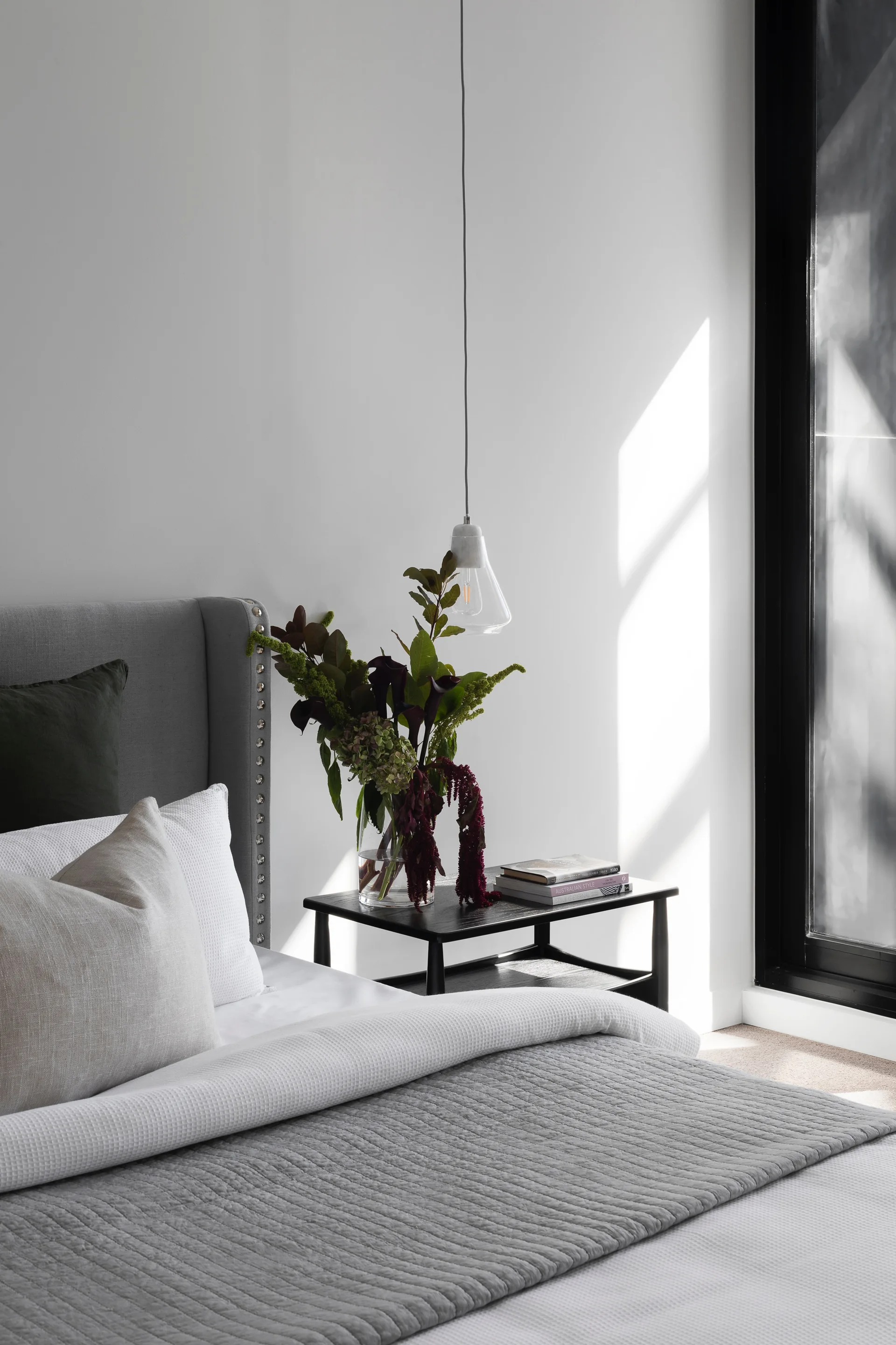 A modern bedroom featuring an upholstered grey headboard, a black wooden nightstand with a tall floral vase, and a hanging clear glass pendant light.