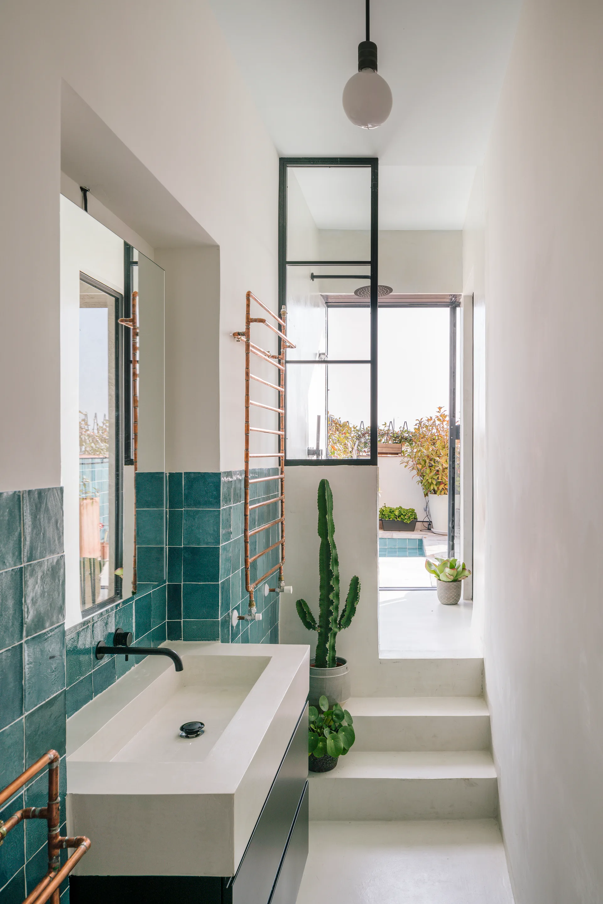 A narrow white bathroom features a concrete trough sink with a black faucet, glossy teal square wall tiles, and white stairs leading to an outdoor terrace.